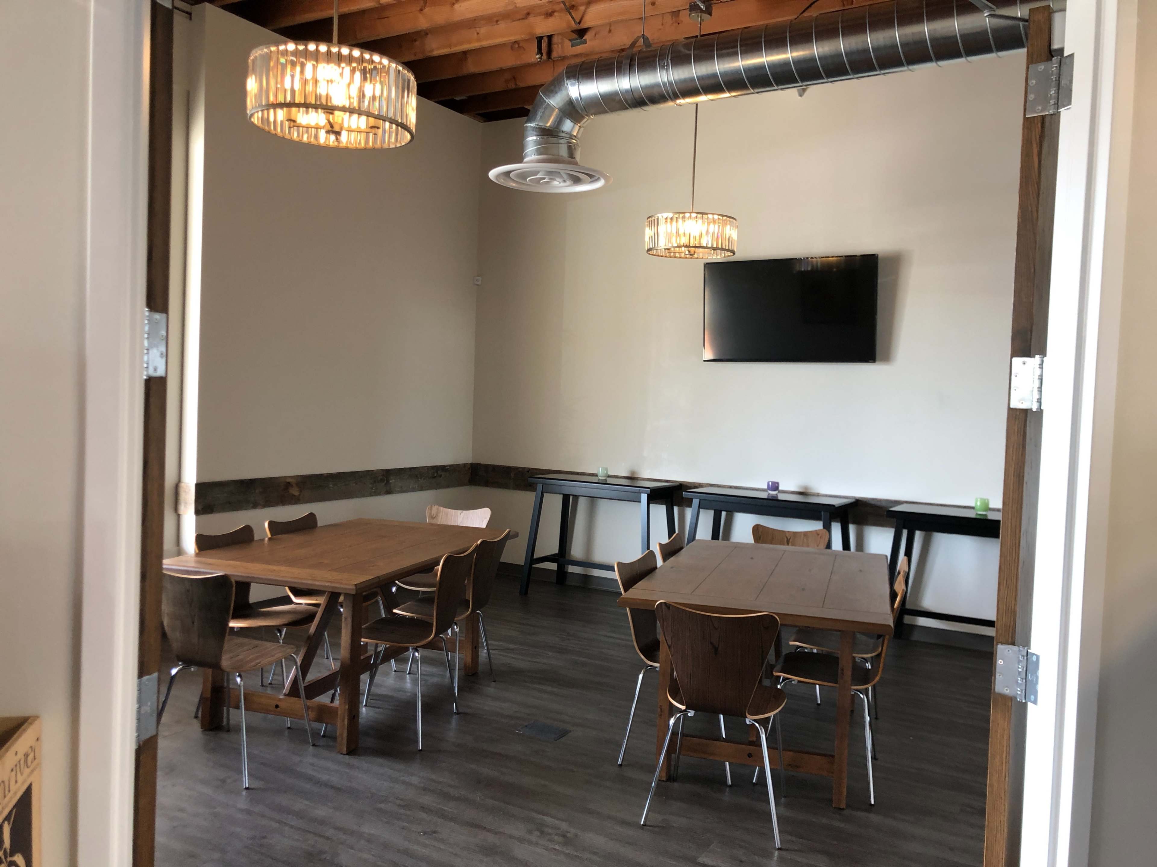 A small, minimalist meeting room with several wooden tables and chairs, two light fixtures hanging from the ceiling, and a wall-mounted television.
