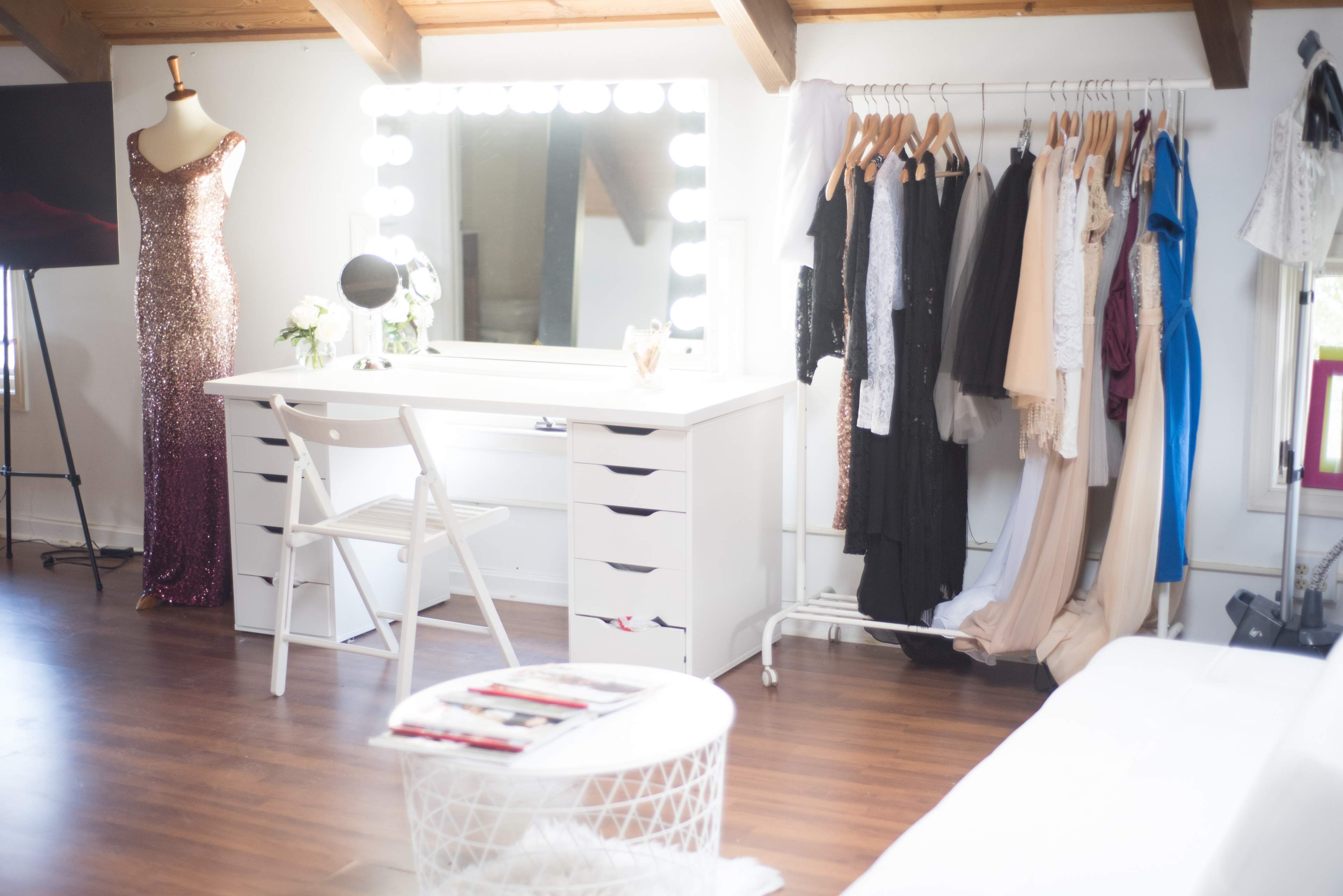 The room features a vanity table with a large illuminated mirror, surrounded by hanging dresses on a rack and a white chair, with a soft seating area visible.
