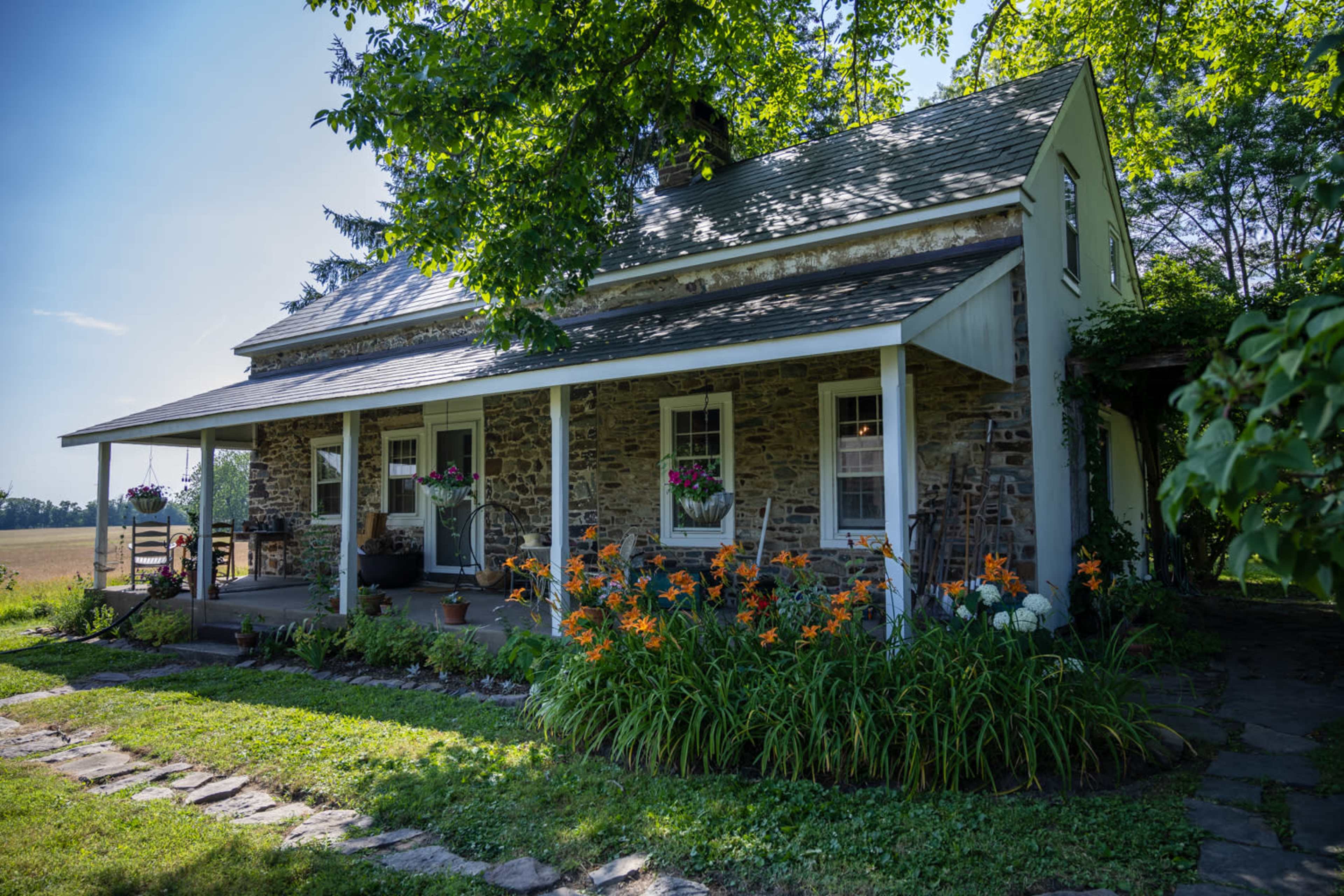 A stone house with a front porch is surrounded by flowers and greenery on a sunny day.