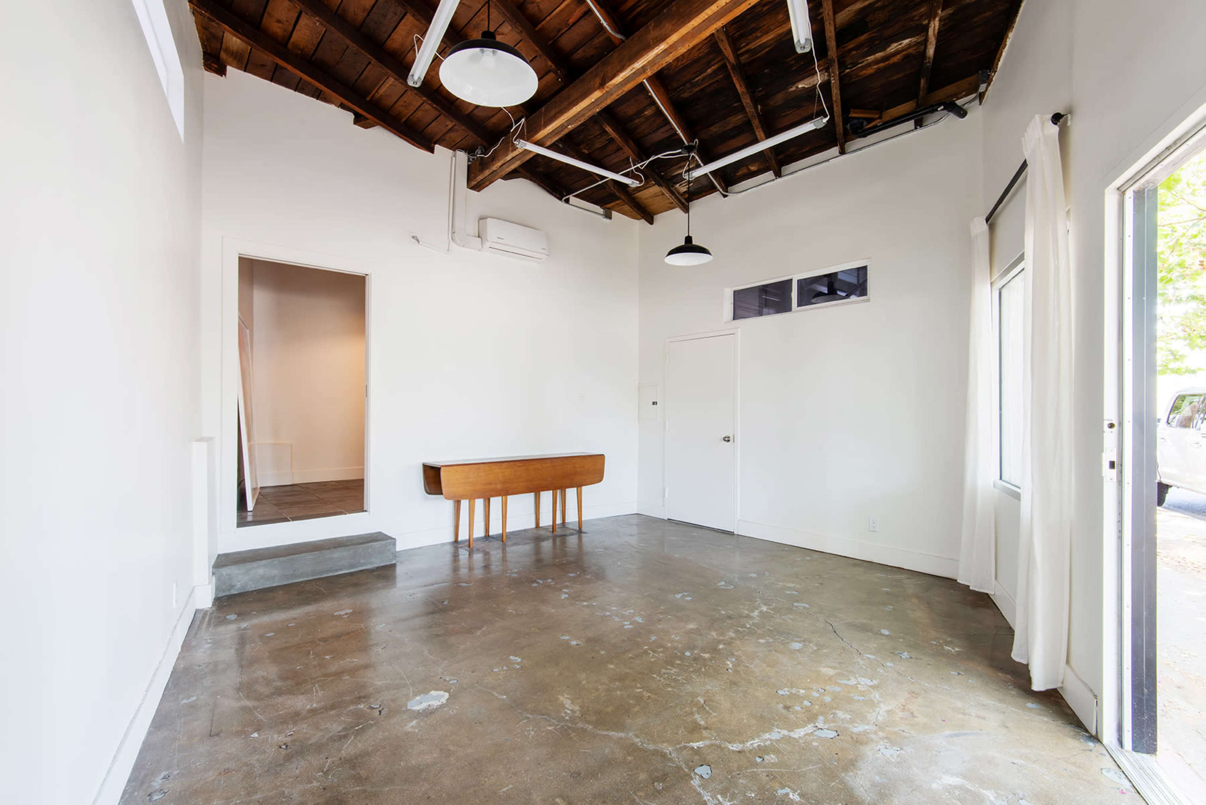 The image shows an empty room with a polished concrete floor, white walls, and exposed wooden beams on the ceiling, featuring a wooden table against one wall and doors leading to an adjoining space and outside.