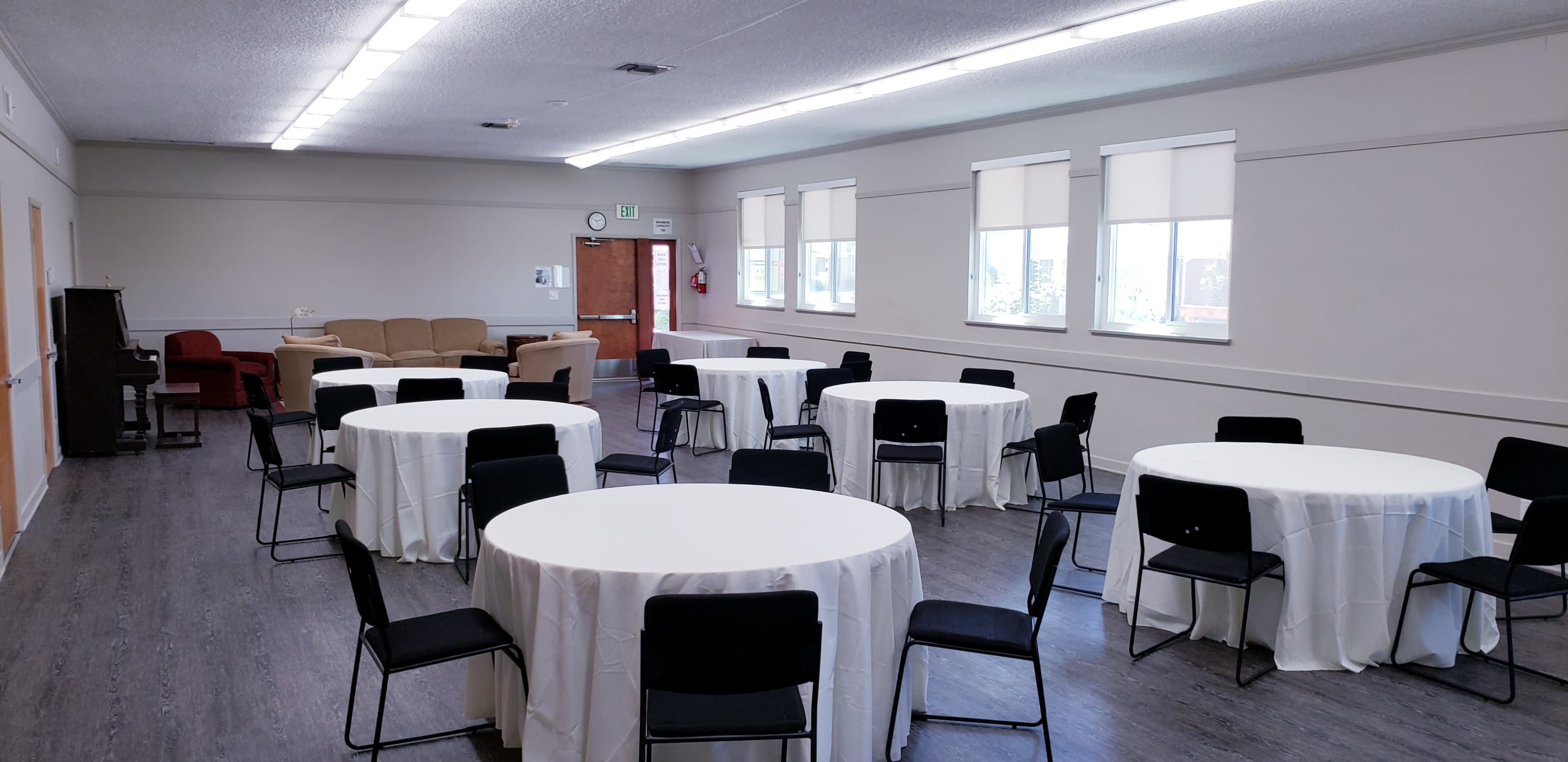 The image shows a spacious room with several round tables covered in white tablecloths and black chairs arranged around them.