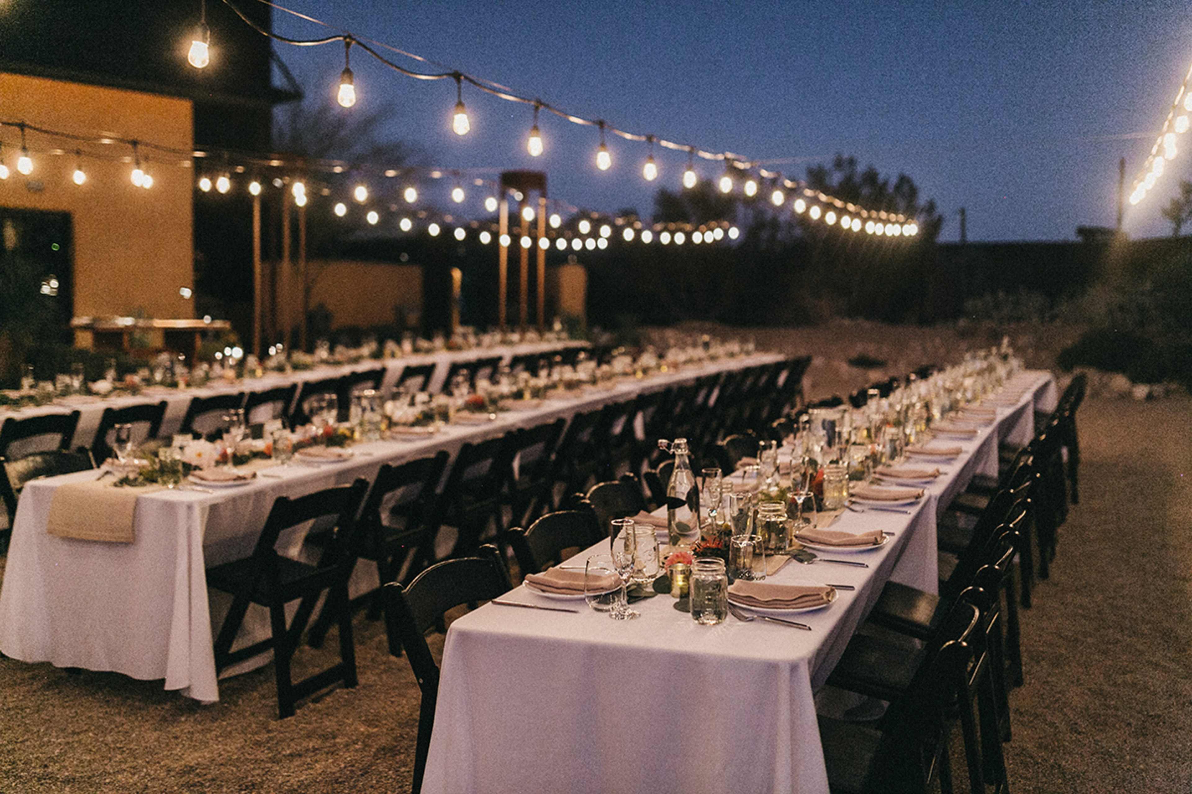 A long outdoor table setup for a dinner event is illuminated by string lights against a twilight sky.