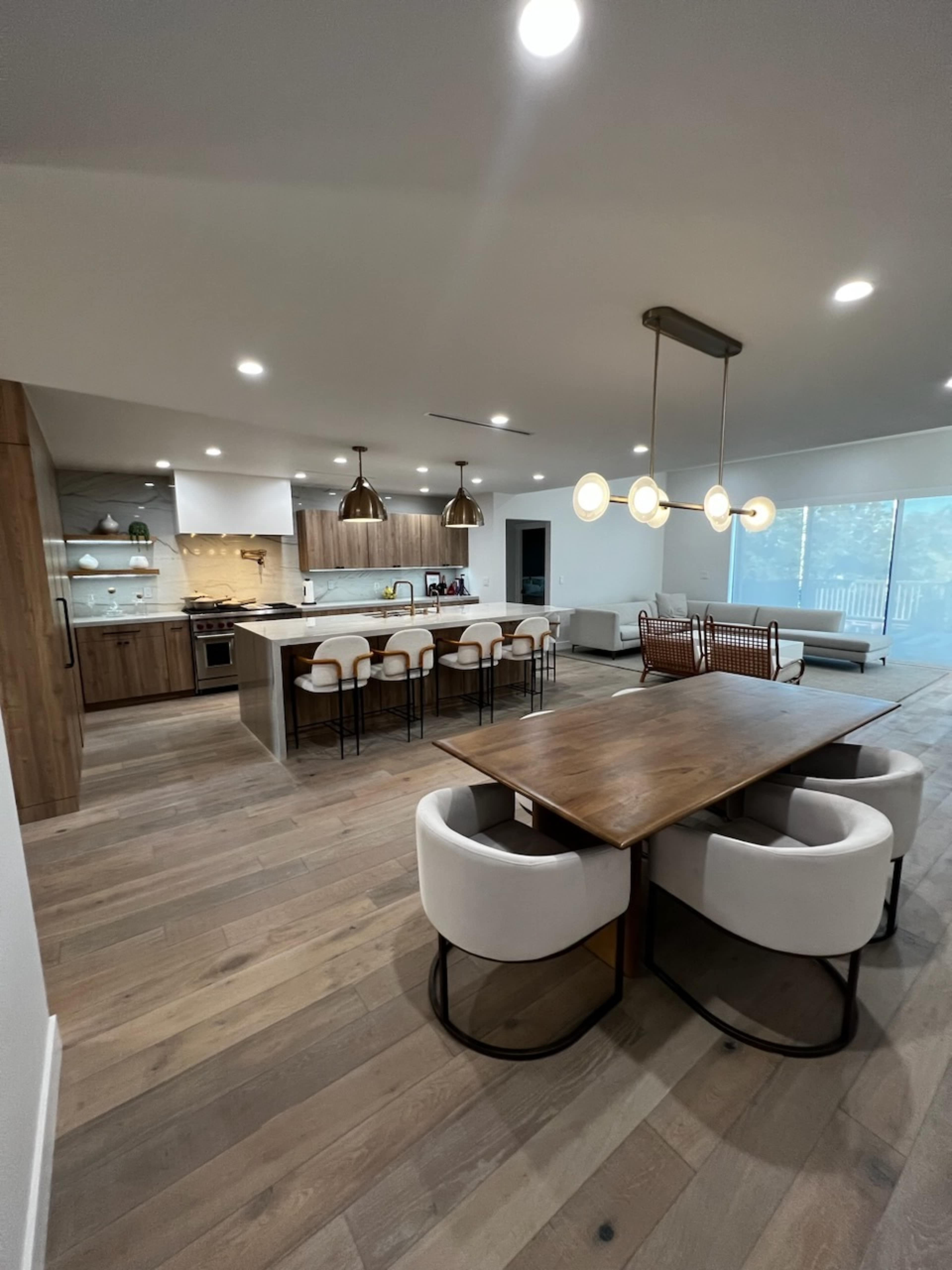 A modern kitchen and dining area with wooden flooring, featuring a large dining table, white upholstered chairs, a kitchen island with bar stools, and stylish pendant lighting.