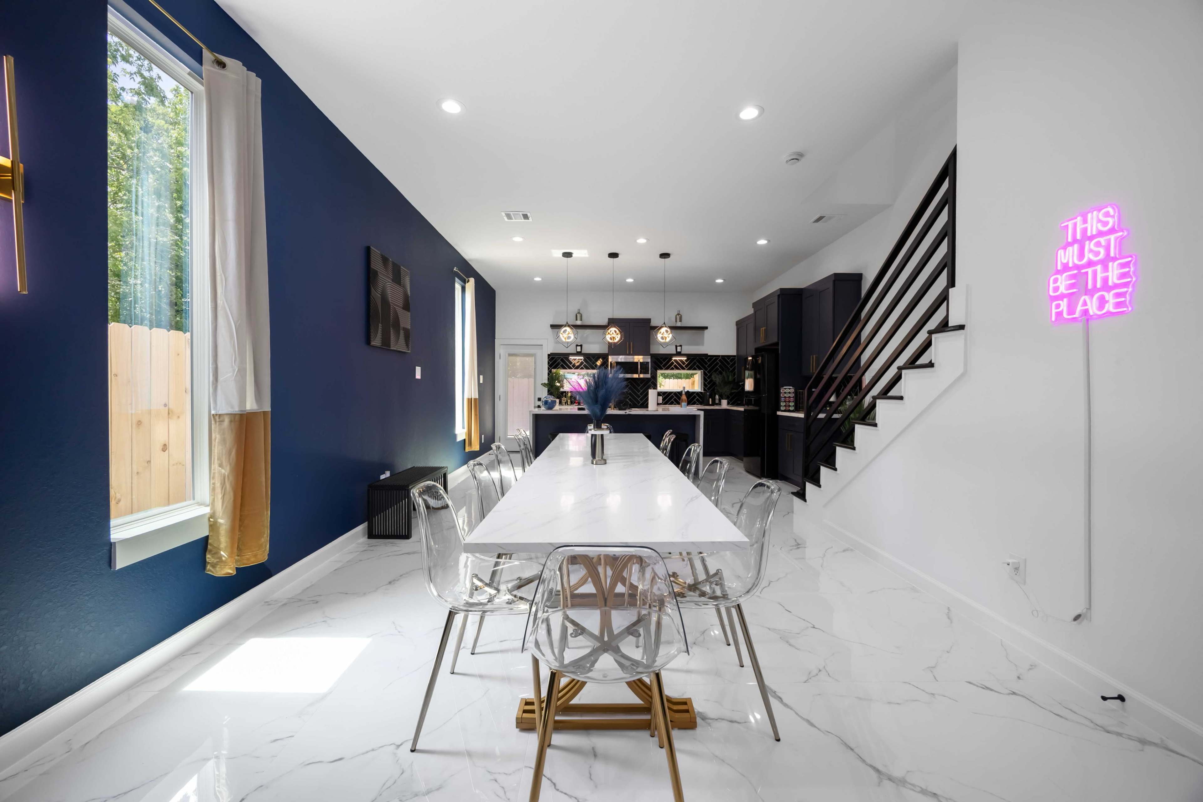 The image shows a modern dining area with a long white table surrounded by clear chairs, leading to a kitchen with dark cabinetry in the background.