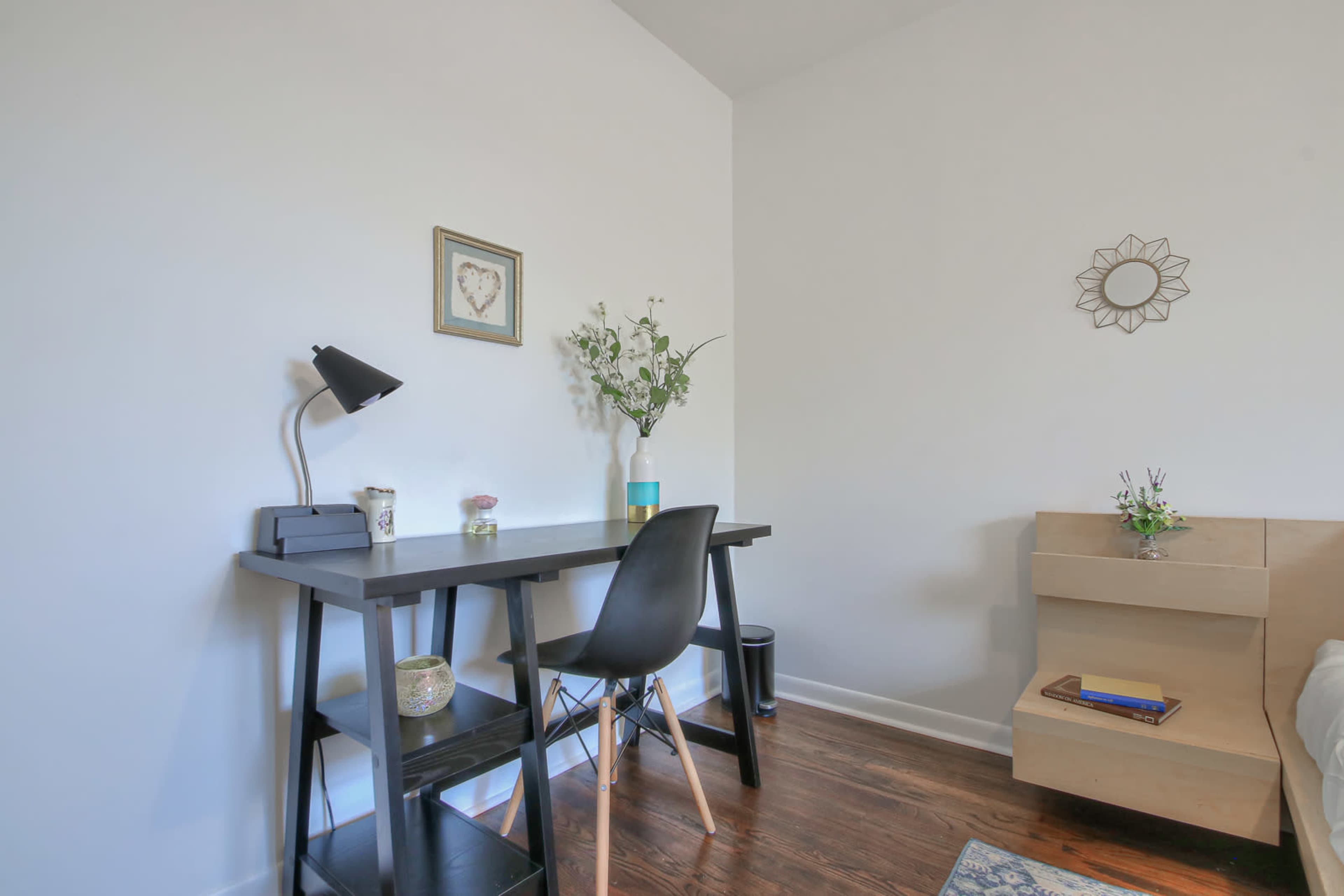 A minimalist workspace features a black desk with a lamp, a vase of flowers, and a wooden chair next to a light-colored bed.