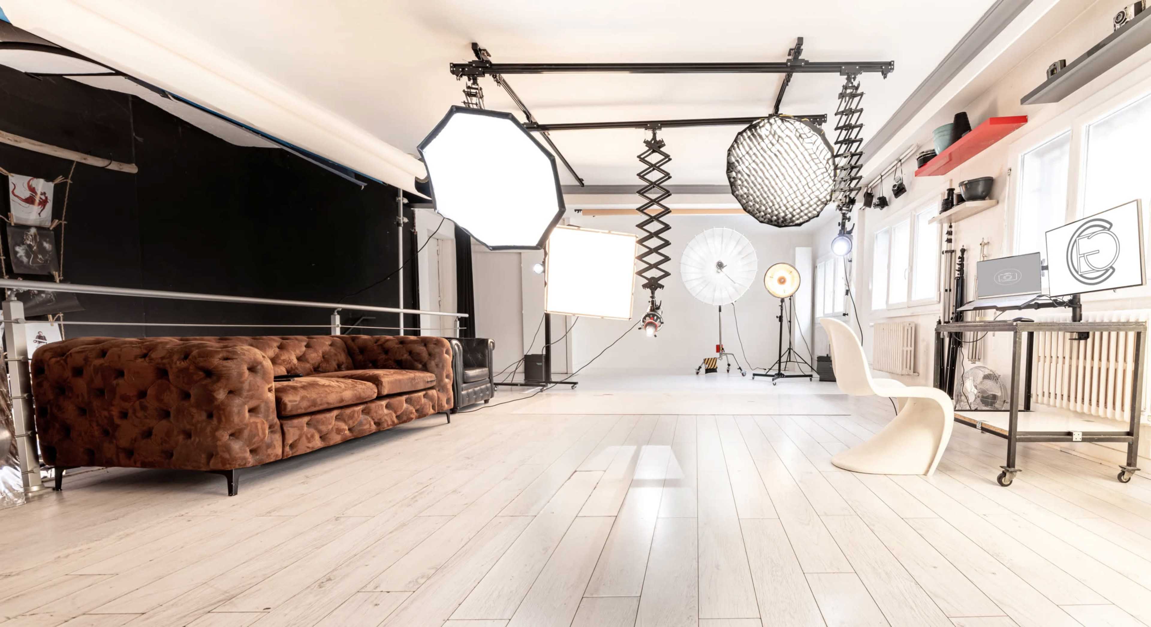 The image shows a photography studio featuring a brown tufted sofa, various light sources, and a minimalist white chair against a backdrop of wooden flooring and a black wall.