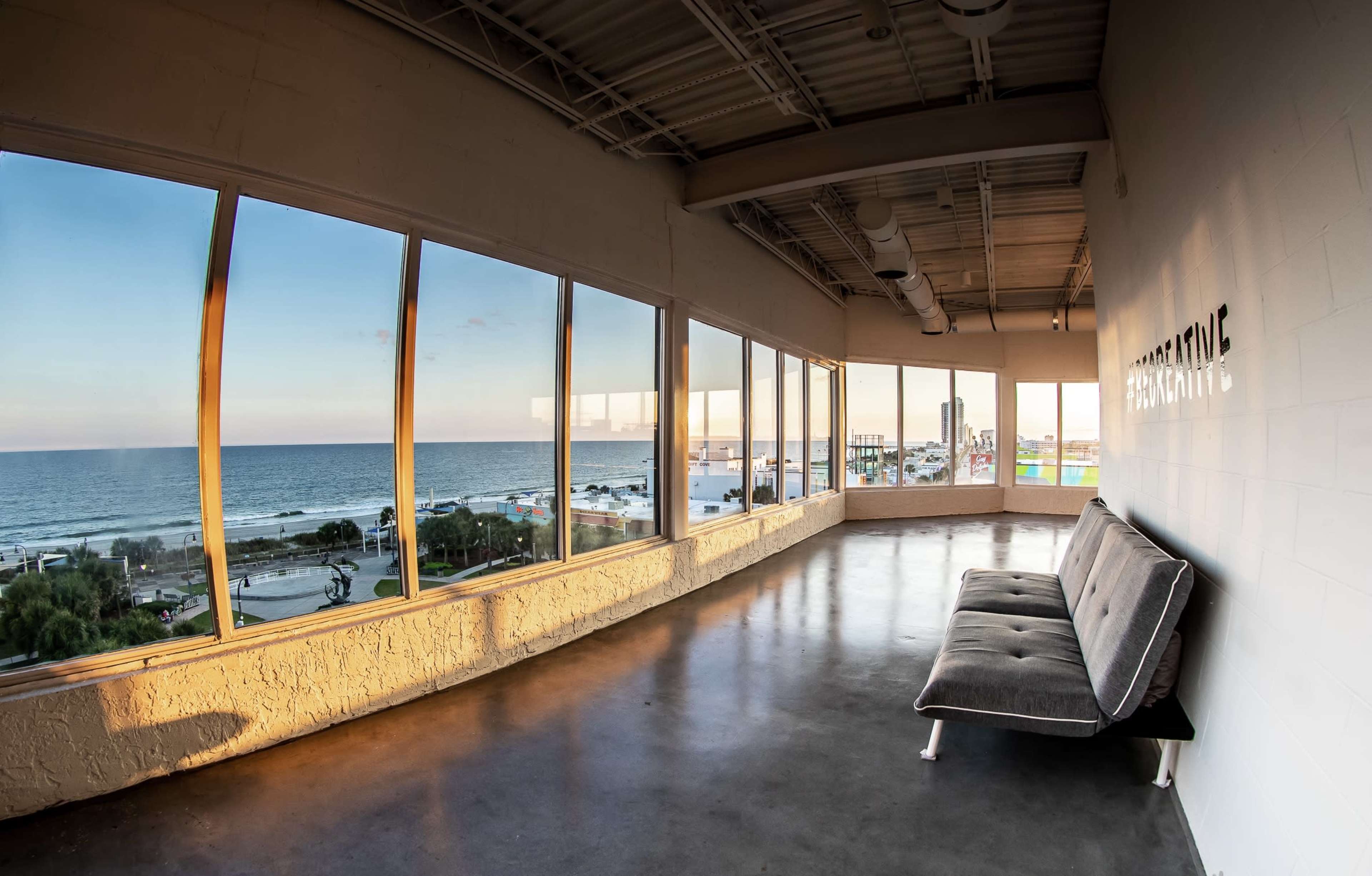 A spacious room with large windows offers a view of the ocean and shoreline at sunset.