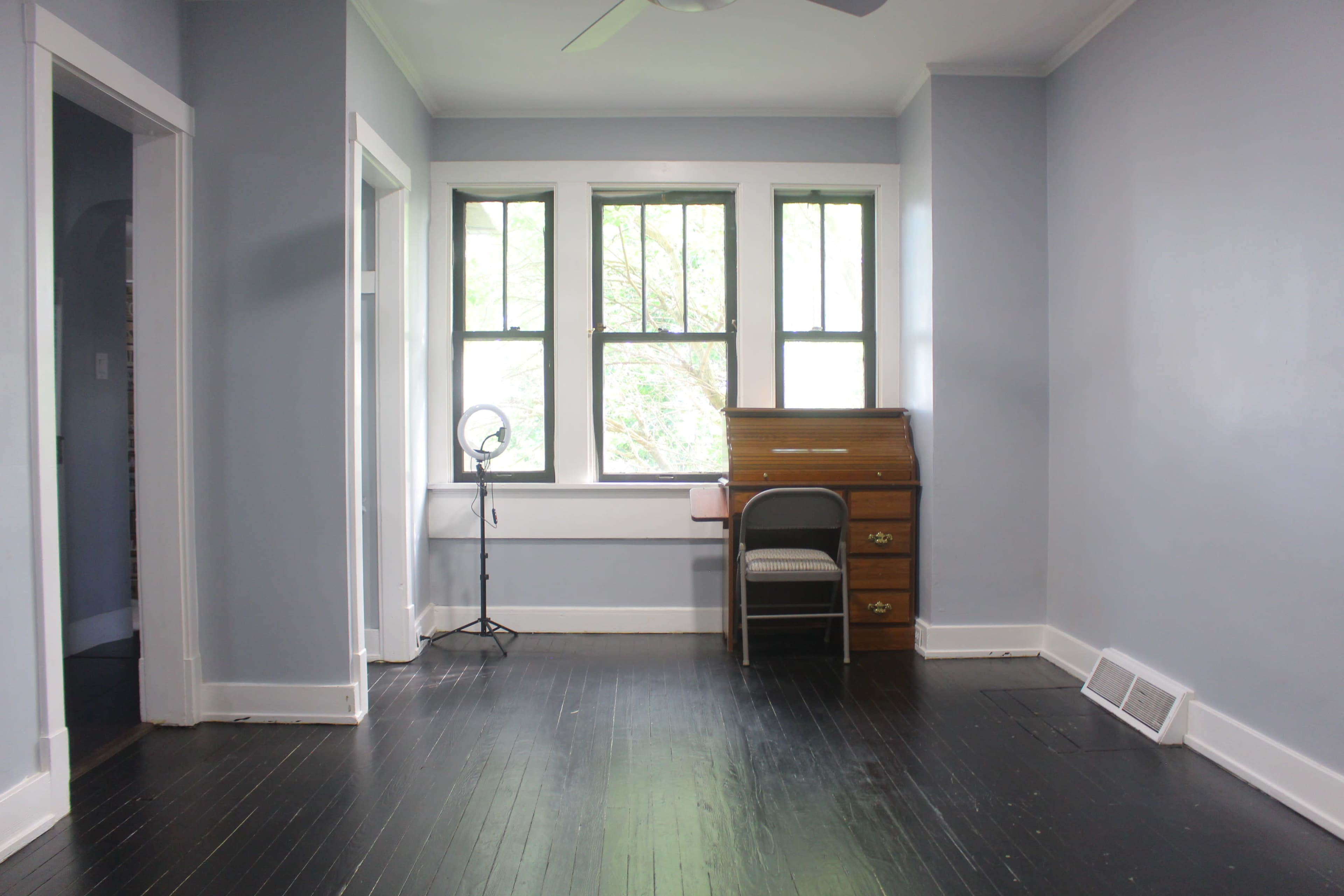 A light blue room features a wooden desk with a chair, a floor lamp, and windows overlooking greenery.