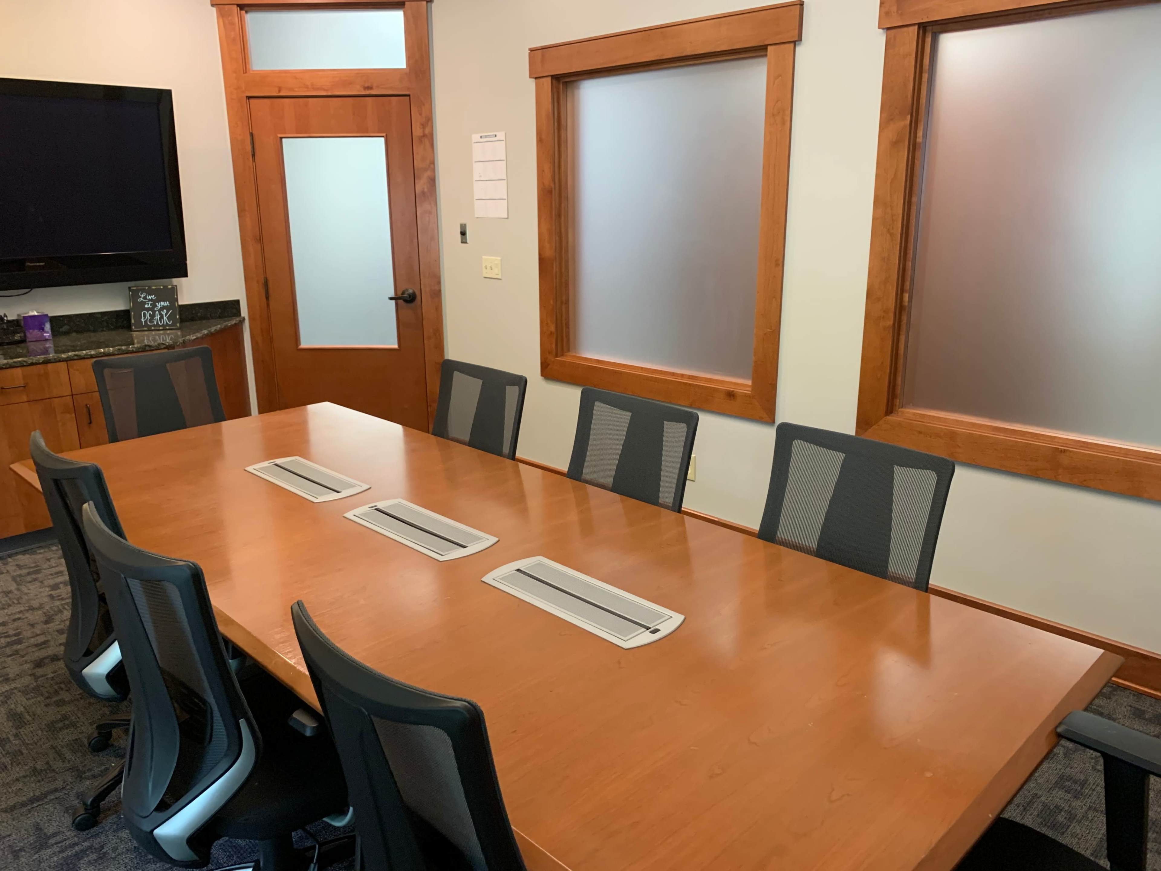 A conference room features a large wooden table surrounded by eight black ergonomic chairs, with frosted glass windows and a television on the wall.