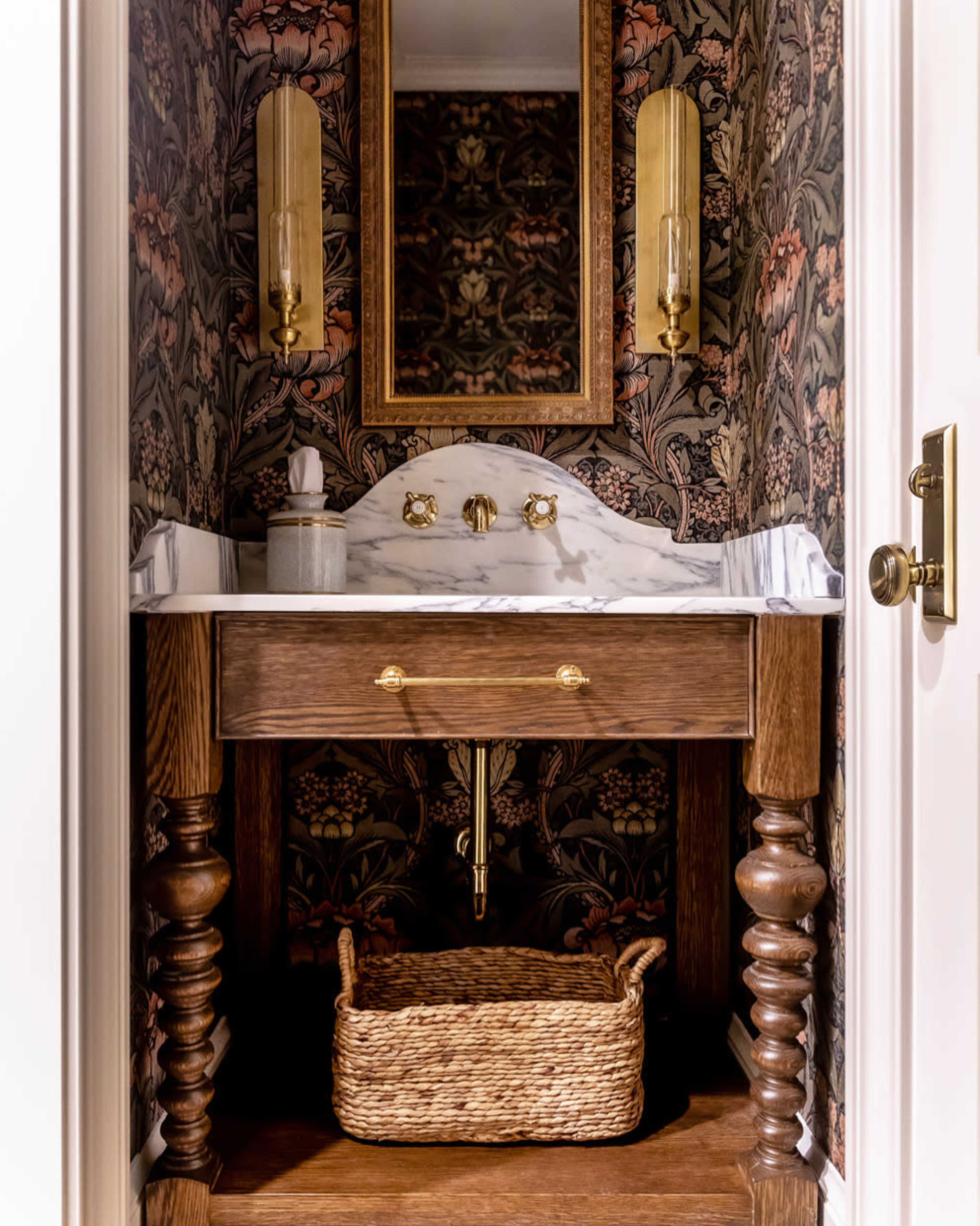 A small bathroom features a wooden vanity with a marble sink, ornate wall wallpaper, and a round mirror above.