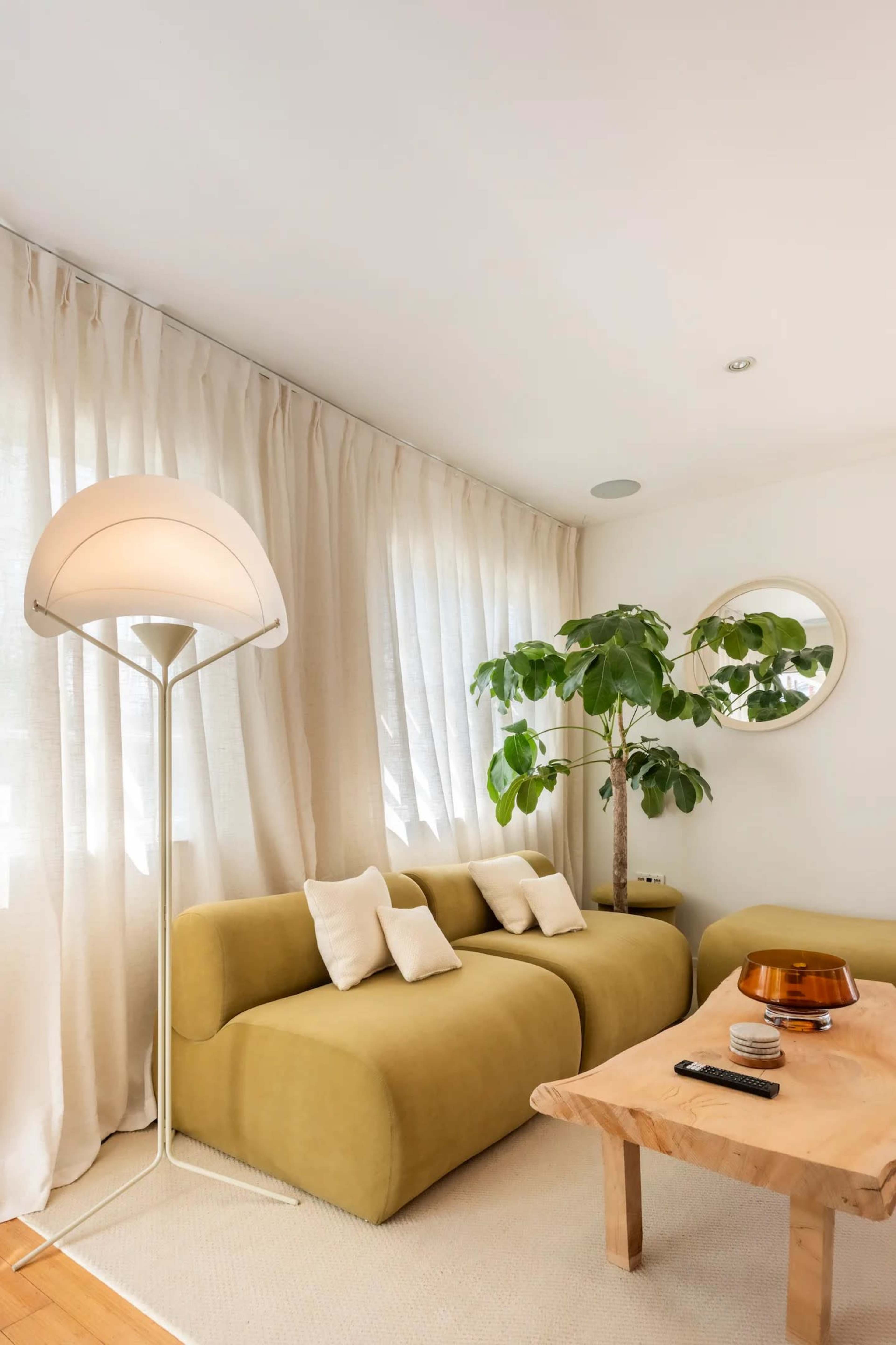 A modern living room features a curved yellow sofa, a wooden coffee table, a potted plant, and large windows dressed with light curtains.
