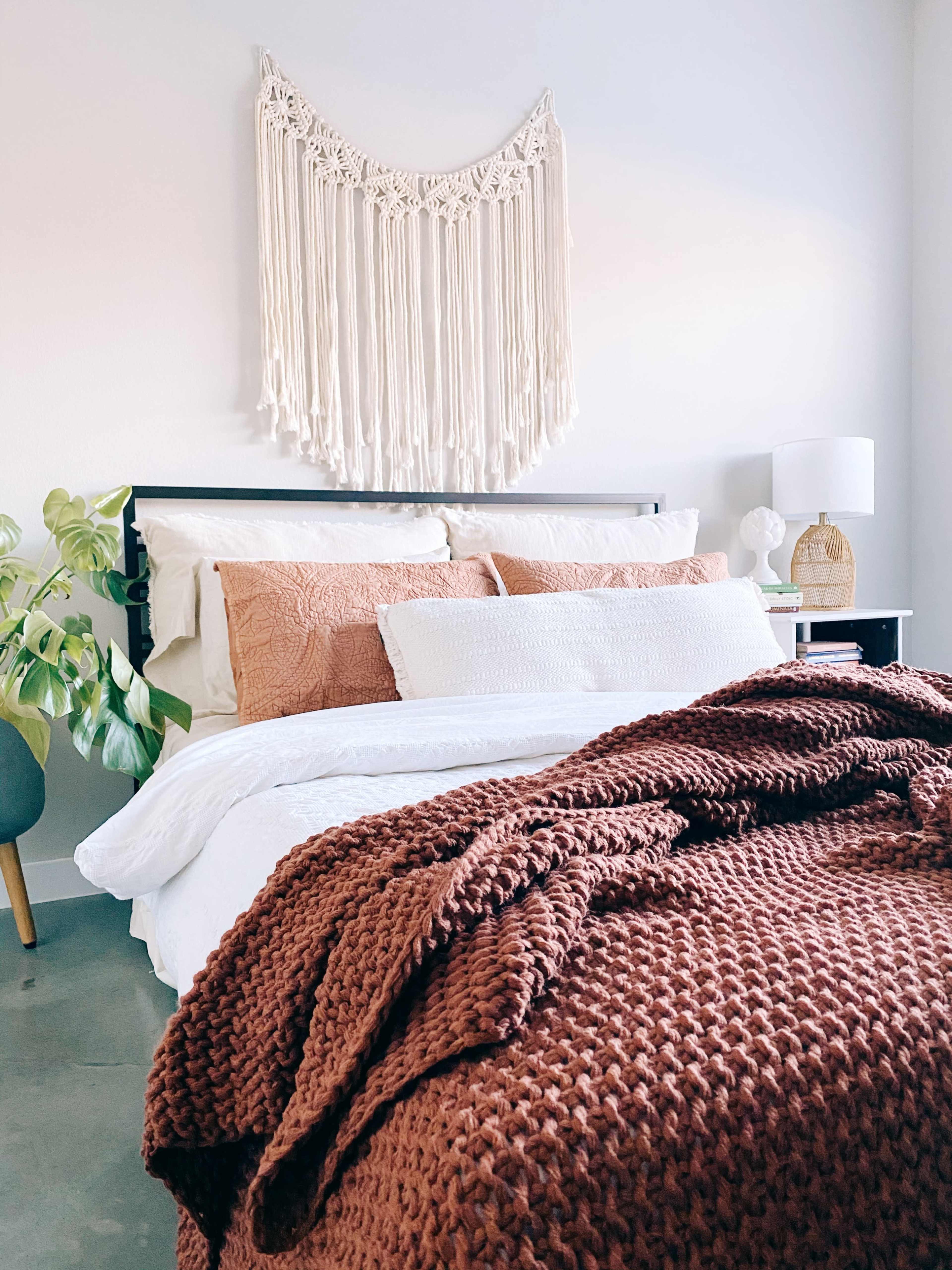 A neatly arranged bed with decorative pillows is set against a light-colored wall, featuring a macramé wall hanging and a bedside lamp.