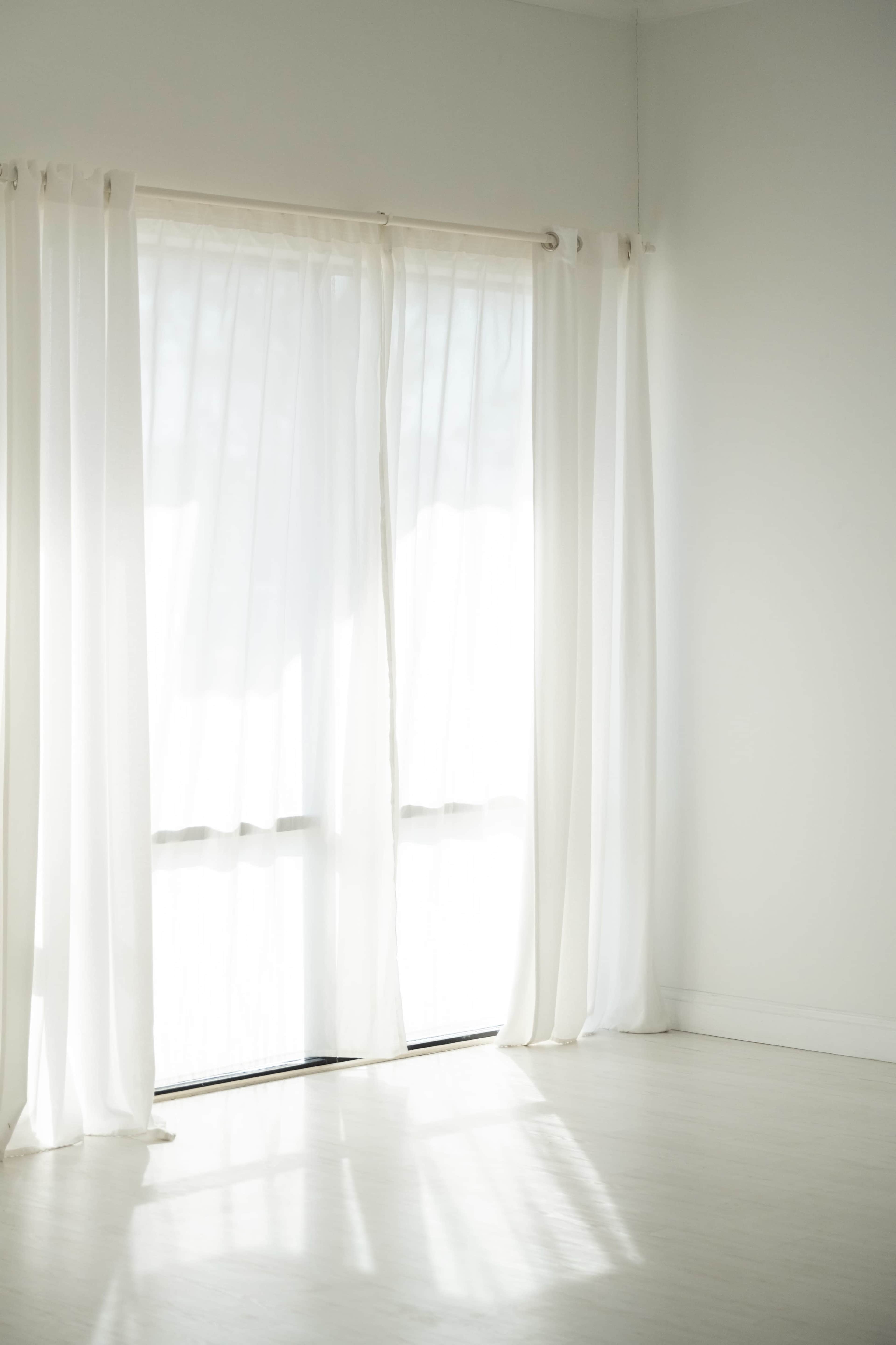The image shows a room with white curtains covering large windows, allowing soft light to filter in, and a pale wooden floor visible beneath.
