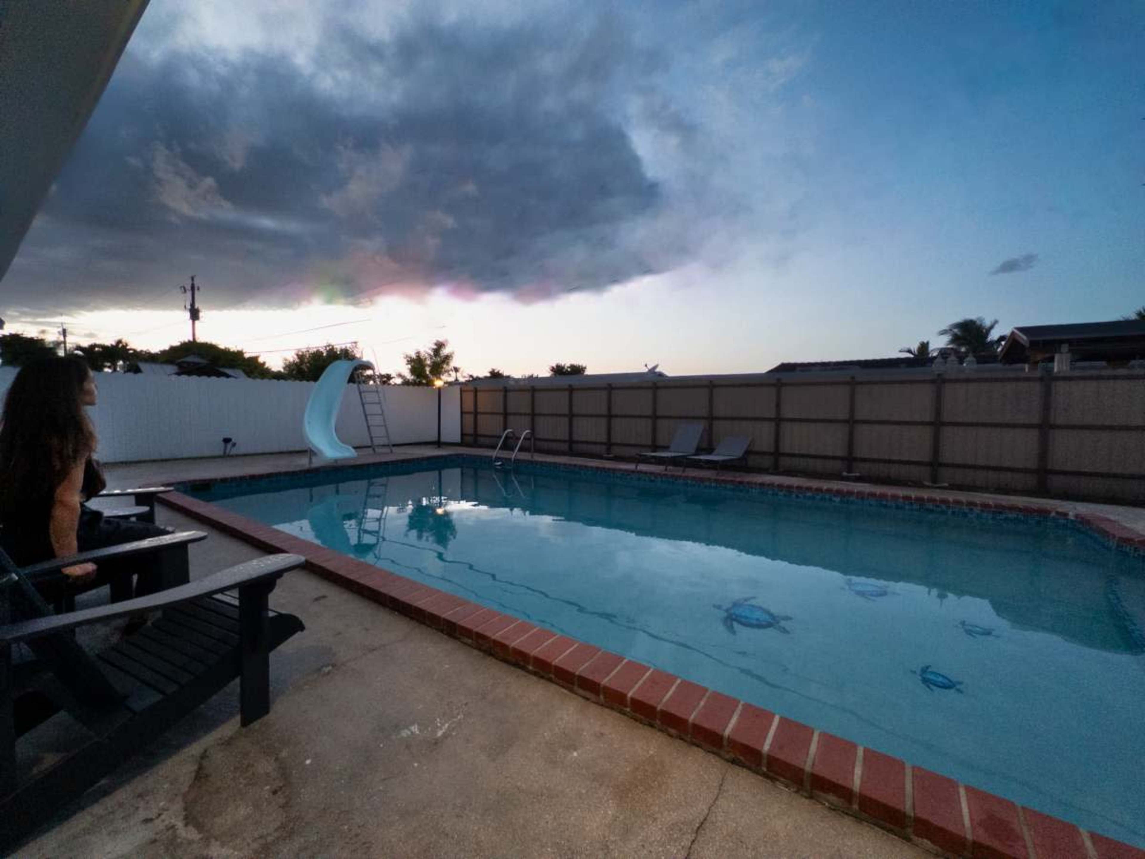 A quiet backyard pool area features a swimming pool with a slide, surrounded by a fence and chairs, under a cloudy evening sky.