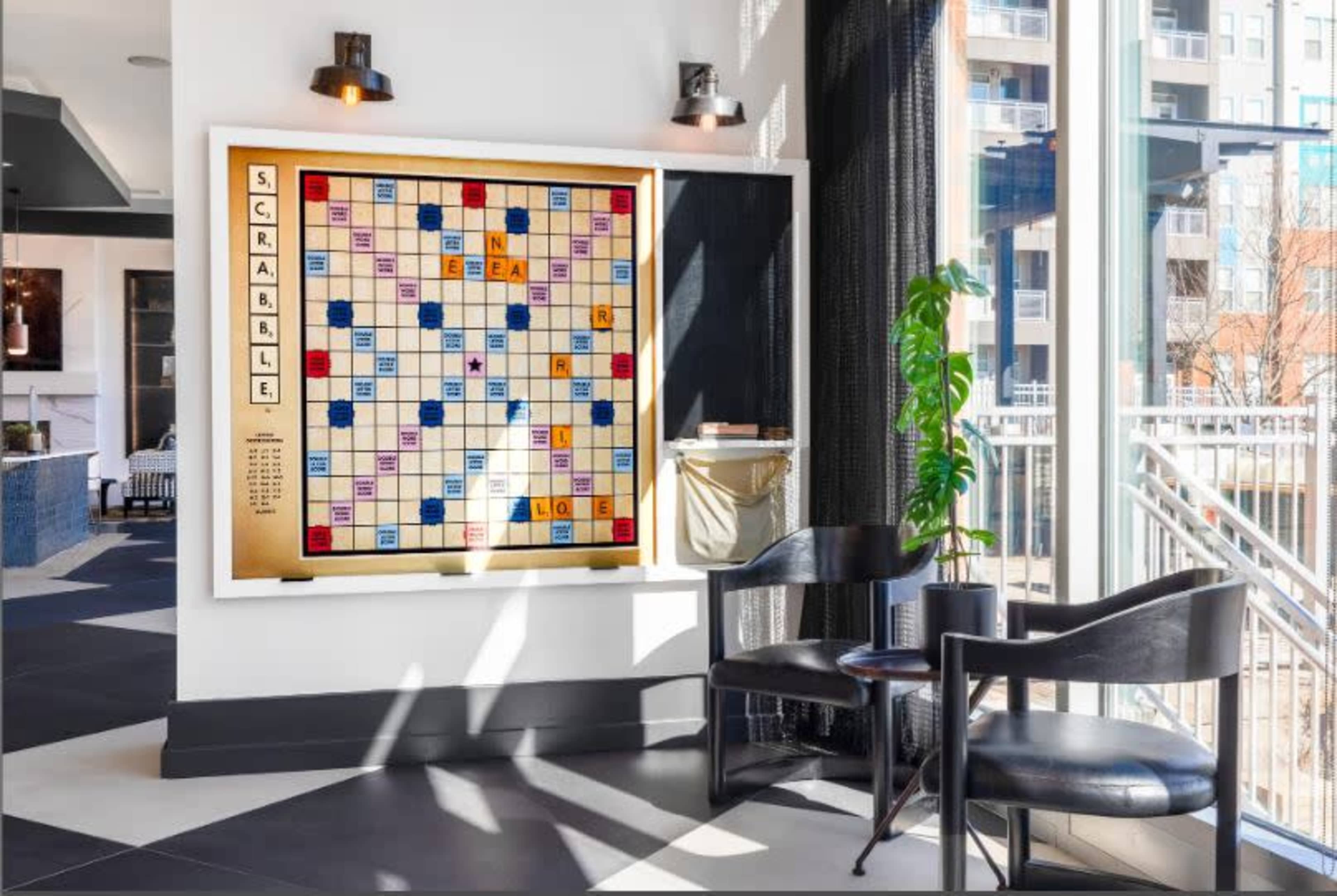 A Scrabble board is displayed on a wall in a modern seating area with large windows, a potted plant, and a black chair.