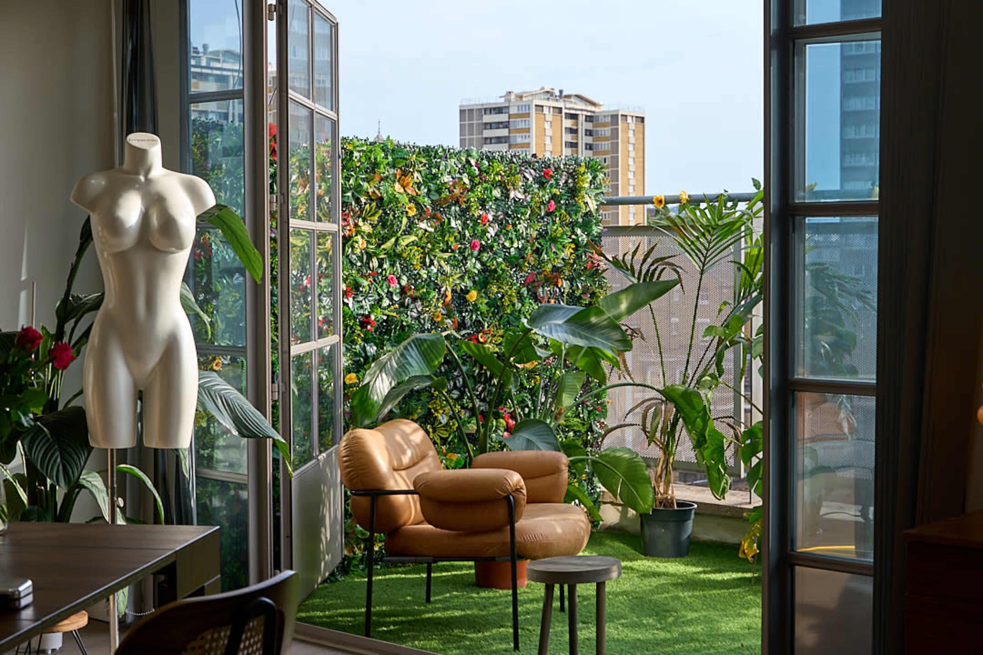 A cozy indoor space features a brown leather chair and a small table, with large windows opening to a balcony adorned with plants and a green trellis of flowers.