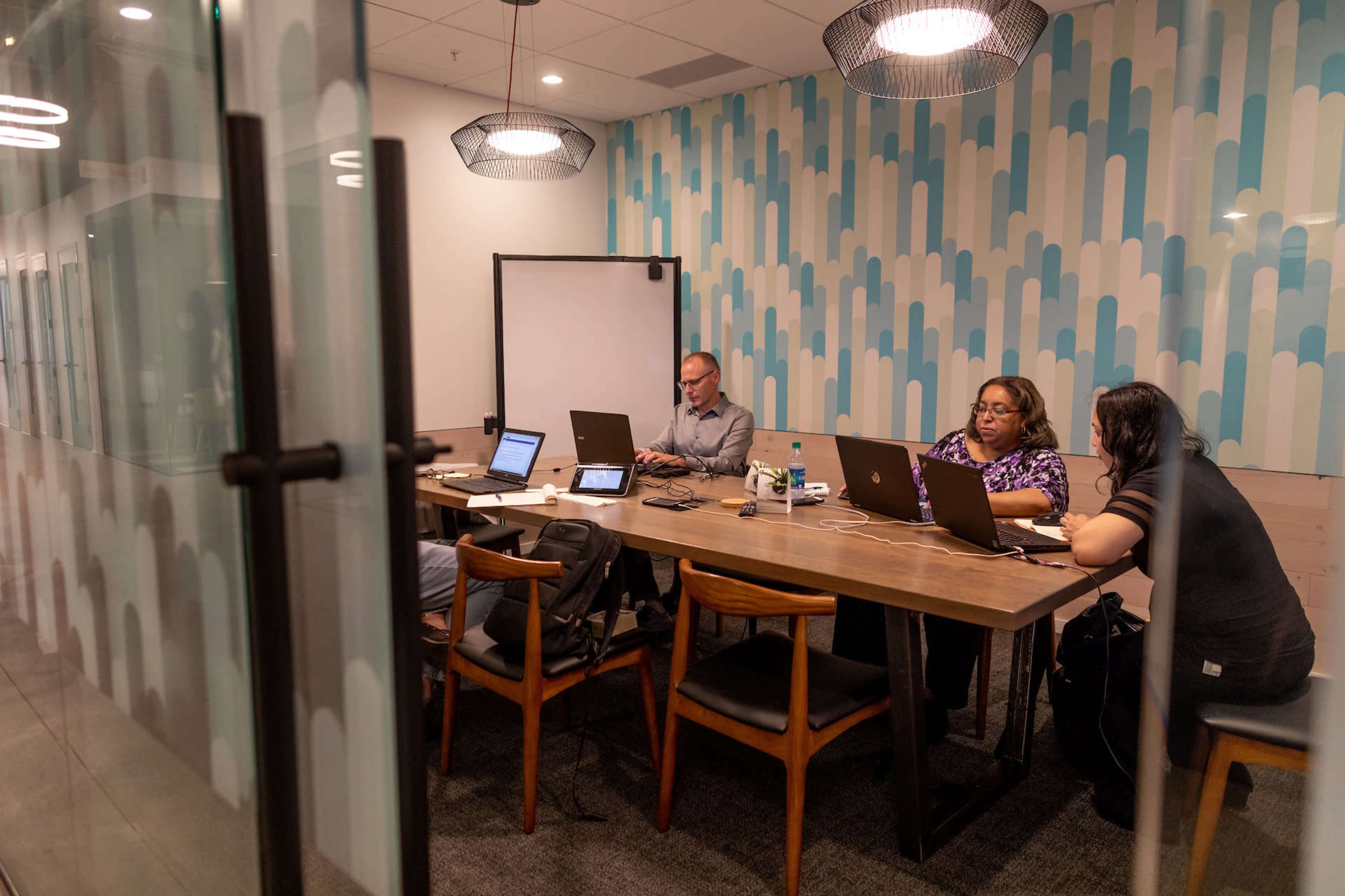 A group of four professionals collaborates at a conference table in a well-lit meeting room, with laptops and tablets in use.