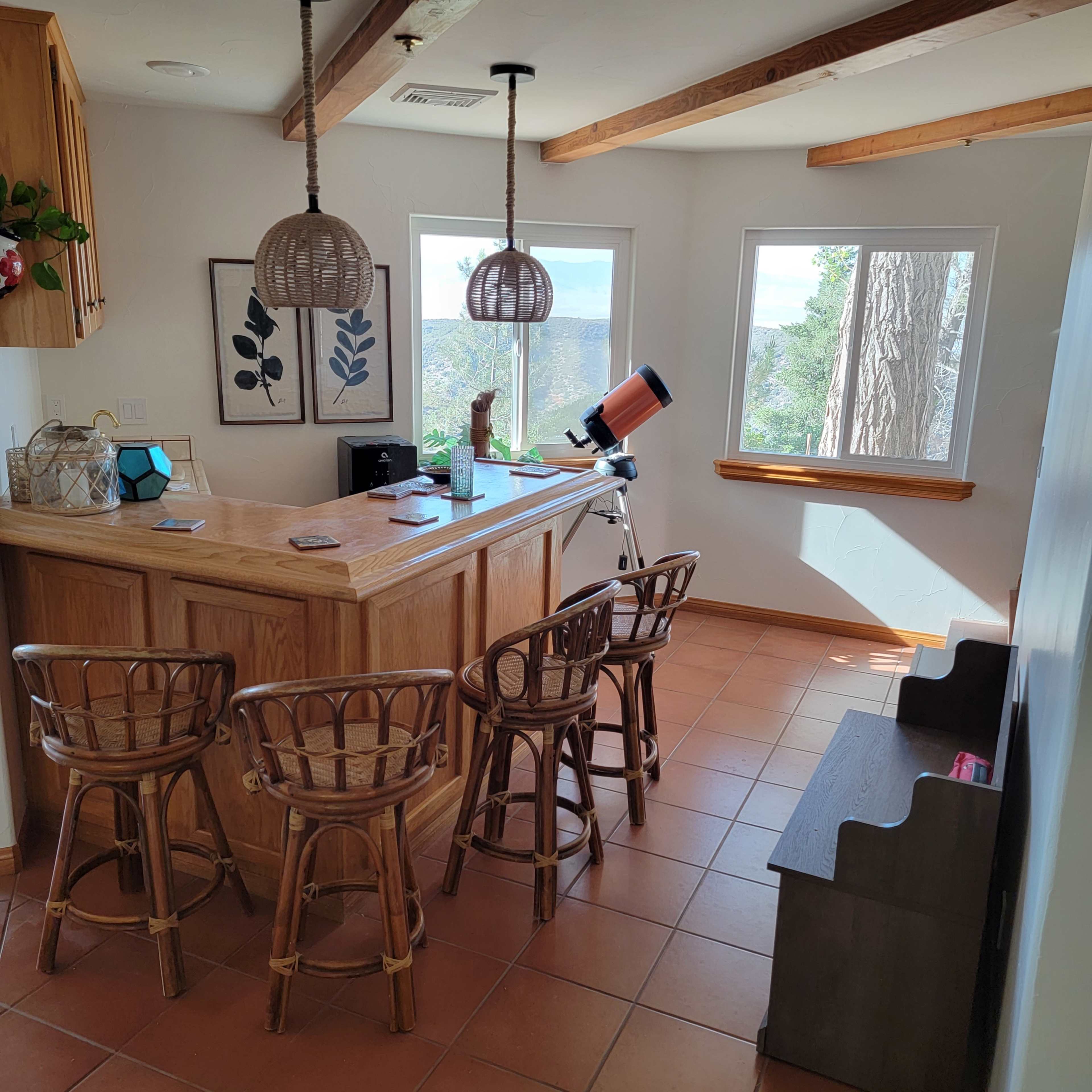 The image shows a sunlit room with a wooden bar and four rattan stools, a telescope positioned by a window overlooking a scenic view.