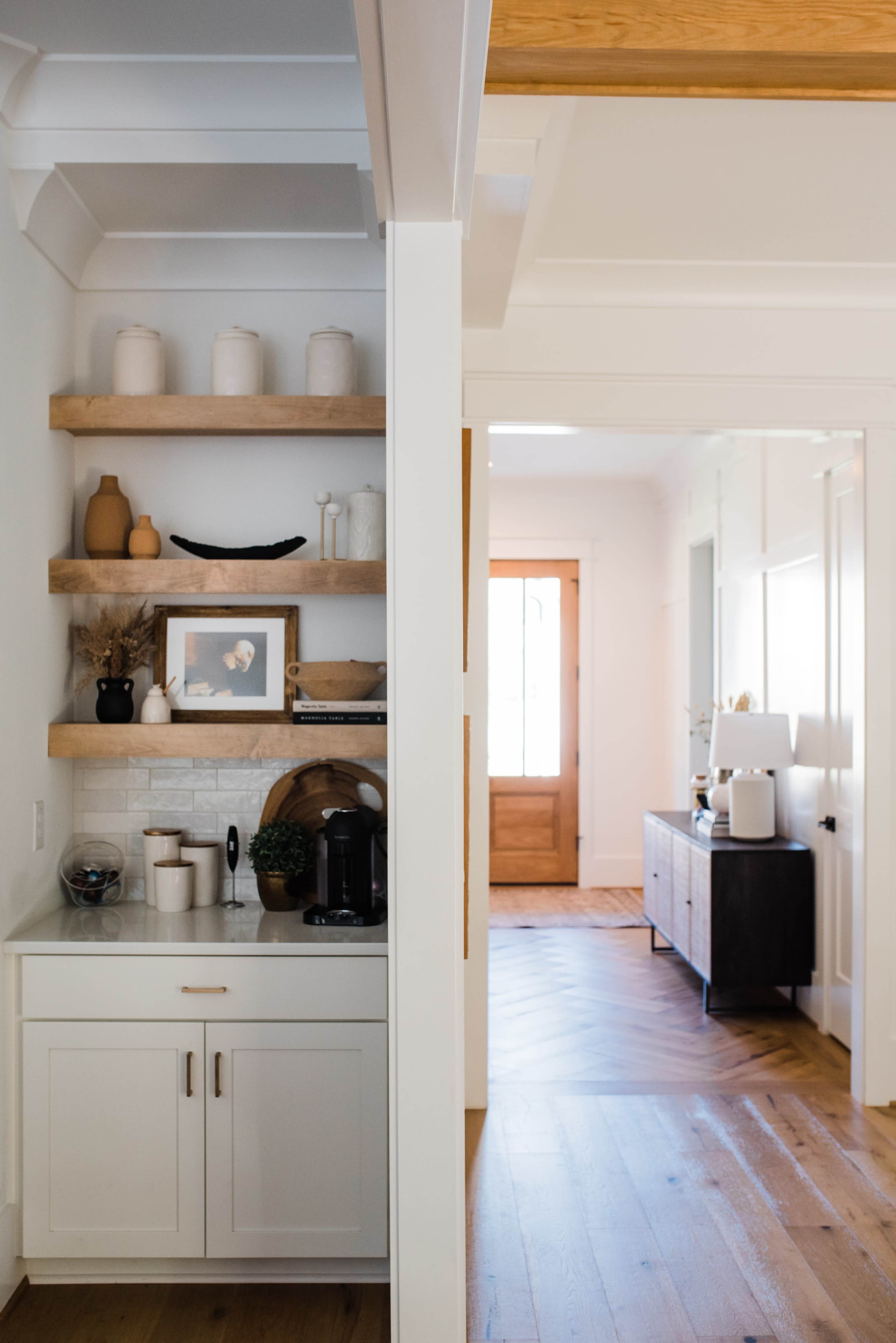 A modern home interior featuring a kitchen nook with open shelves displaying decorative items, while a hallway leads to a wooden front door.