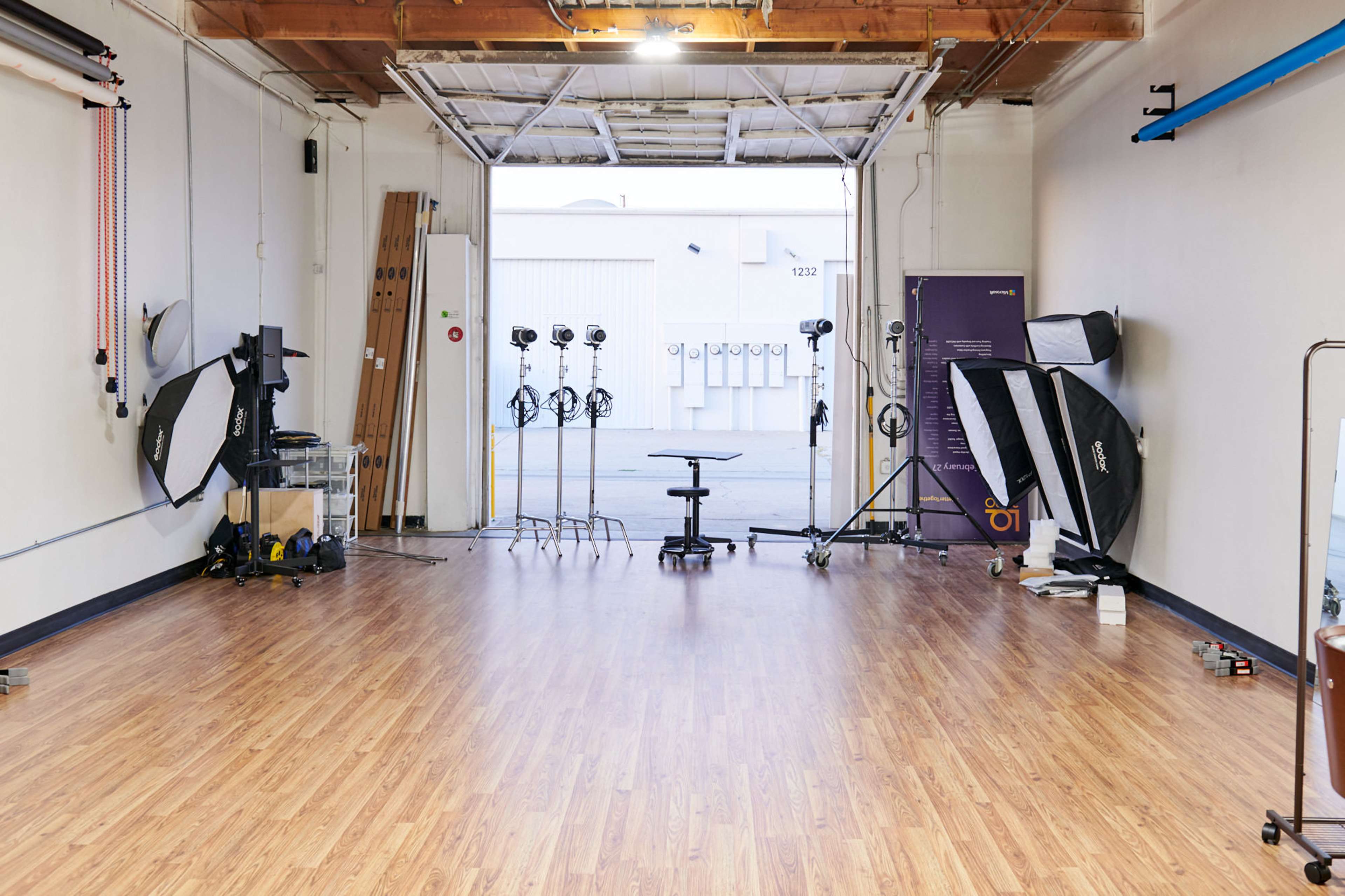 A spacious photography studio with wooden flooring, multiple light stands, softboxes, and a central rolling chair, all set up for a shoot.