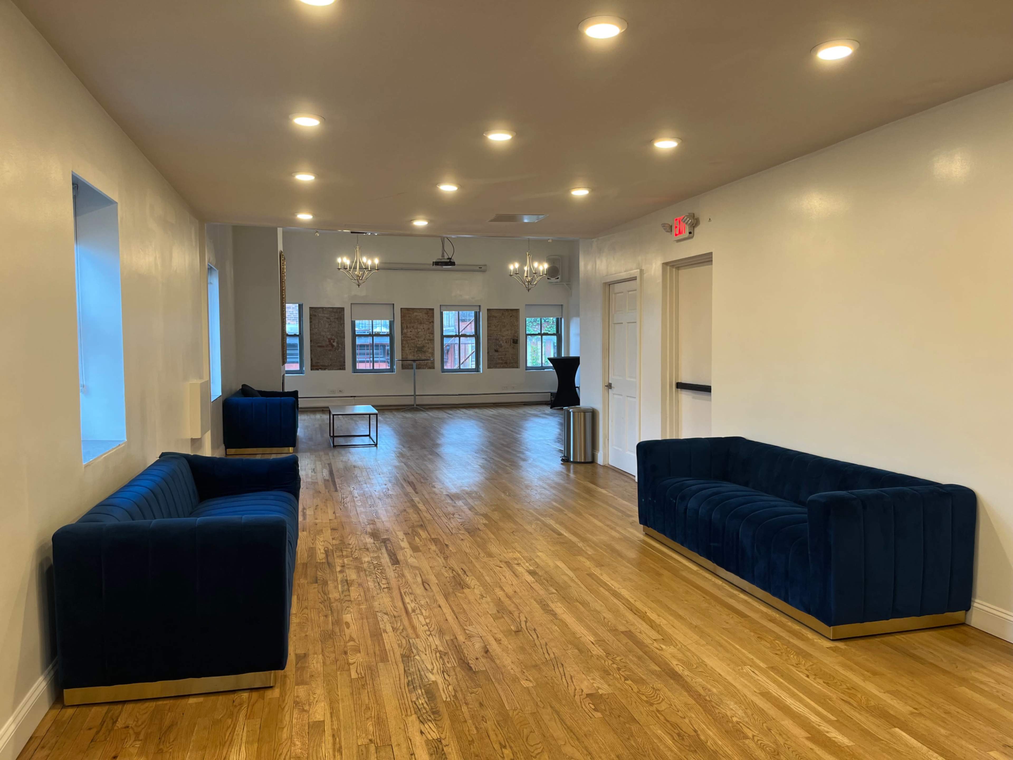 The image shows a well-lit, spacious interior room with wooden floors, two navy blue sofas, a coffee table, and large windows.