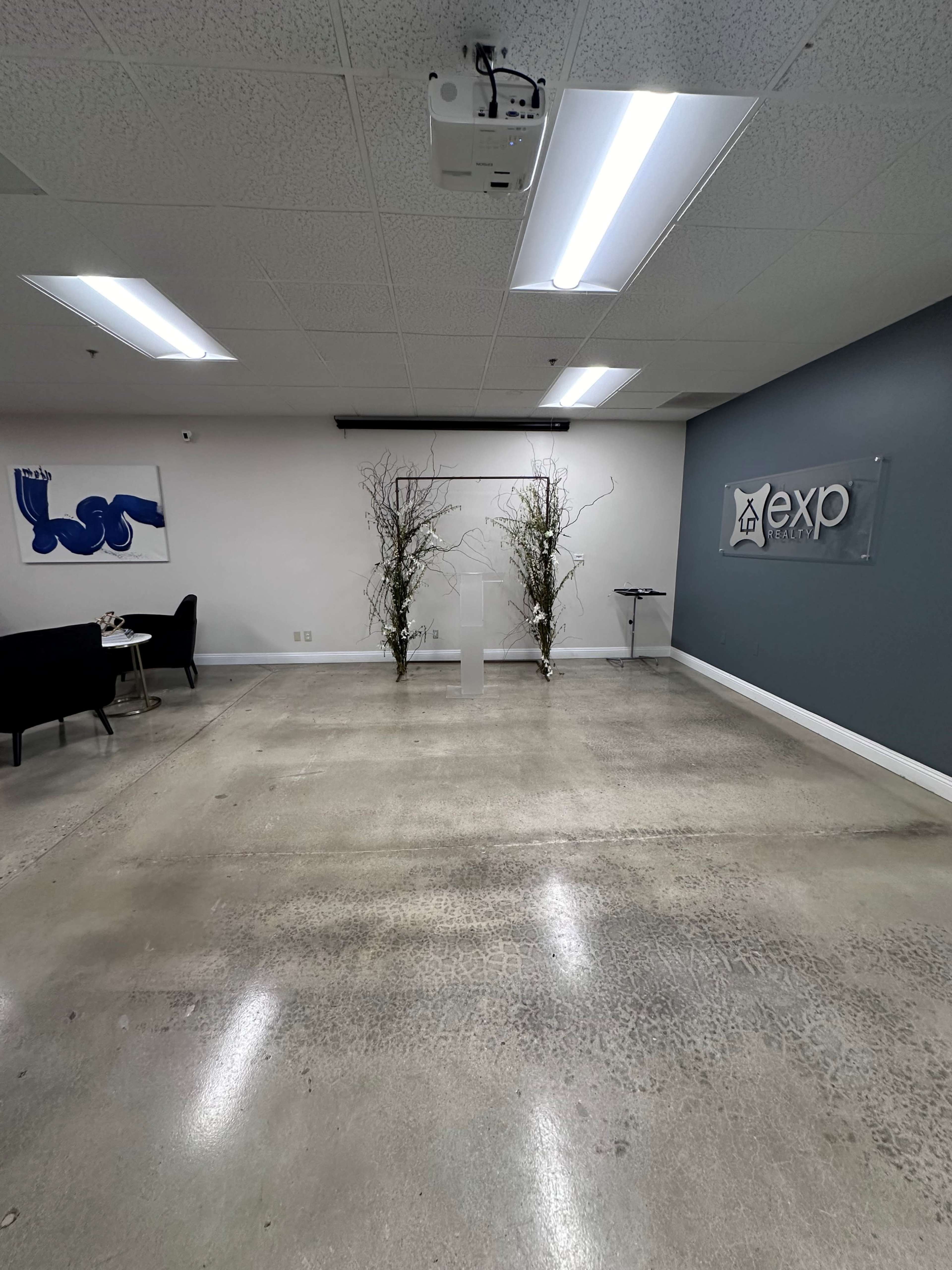 A spacious, modern office interior with a polished concrete floor, featuring decorative branches on either side of a white pedestal and a logo display on the wall.