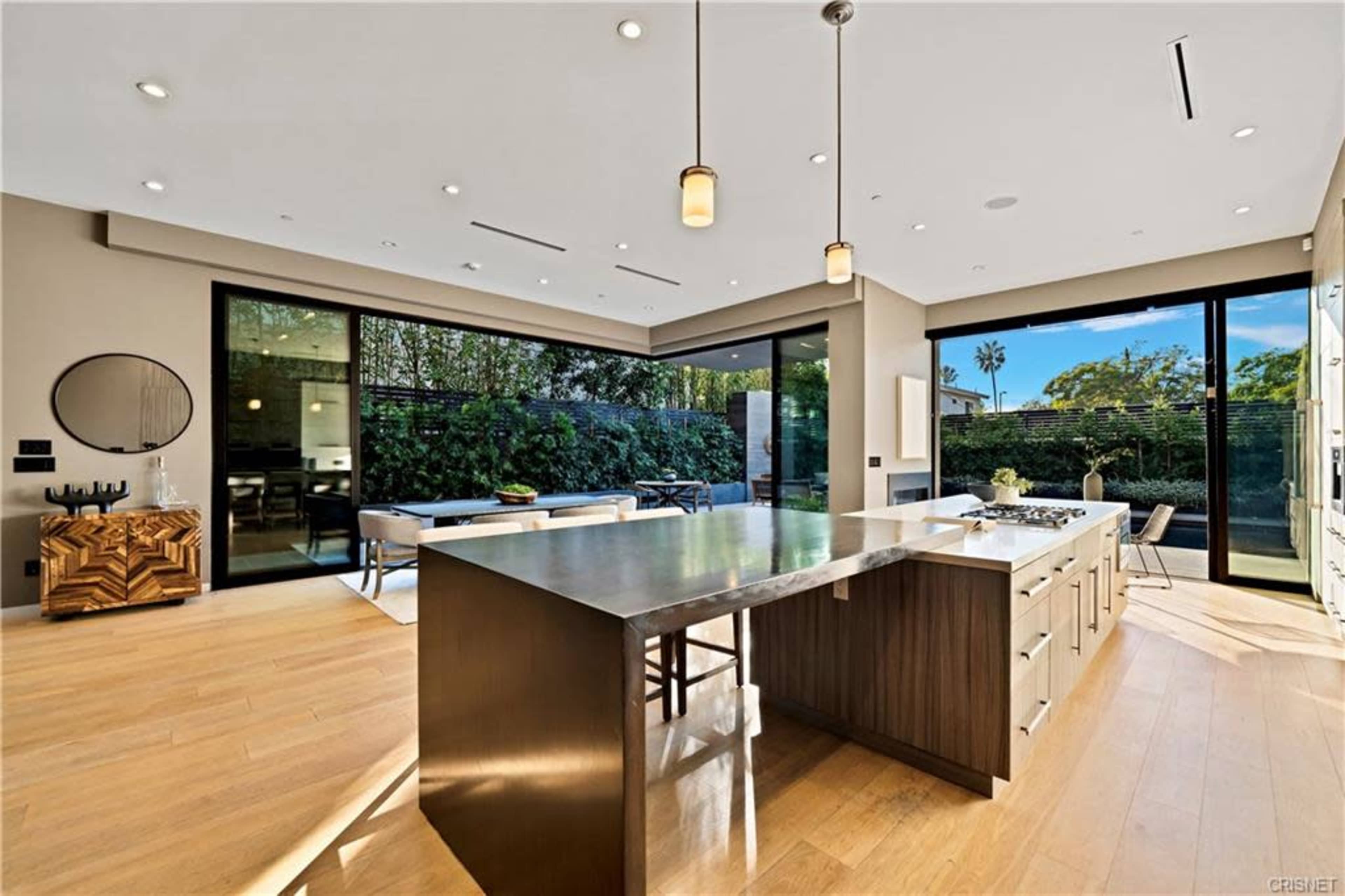 A modern kitchen features a central island with a stainless steel countertop, surrounded by sliding glass doors that open to a garden view.