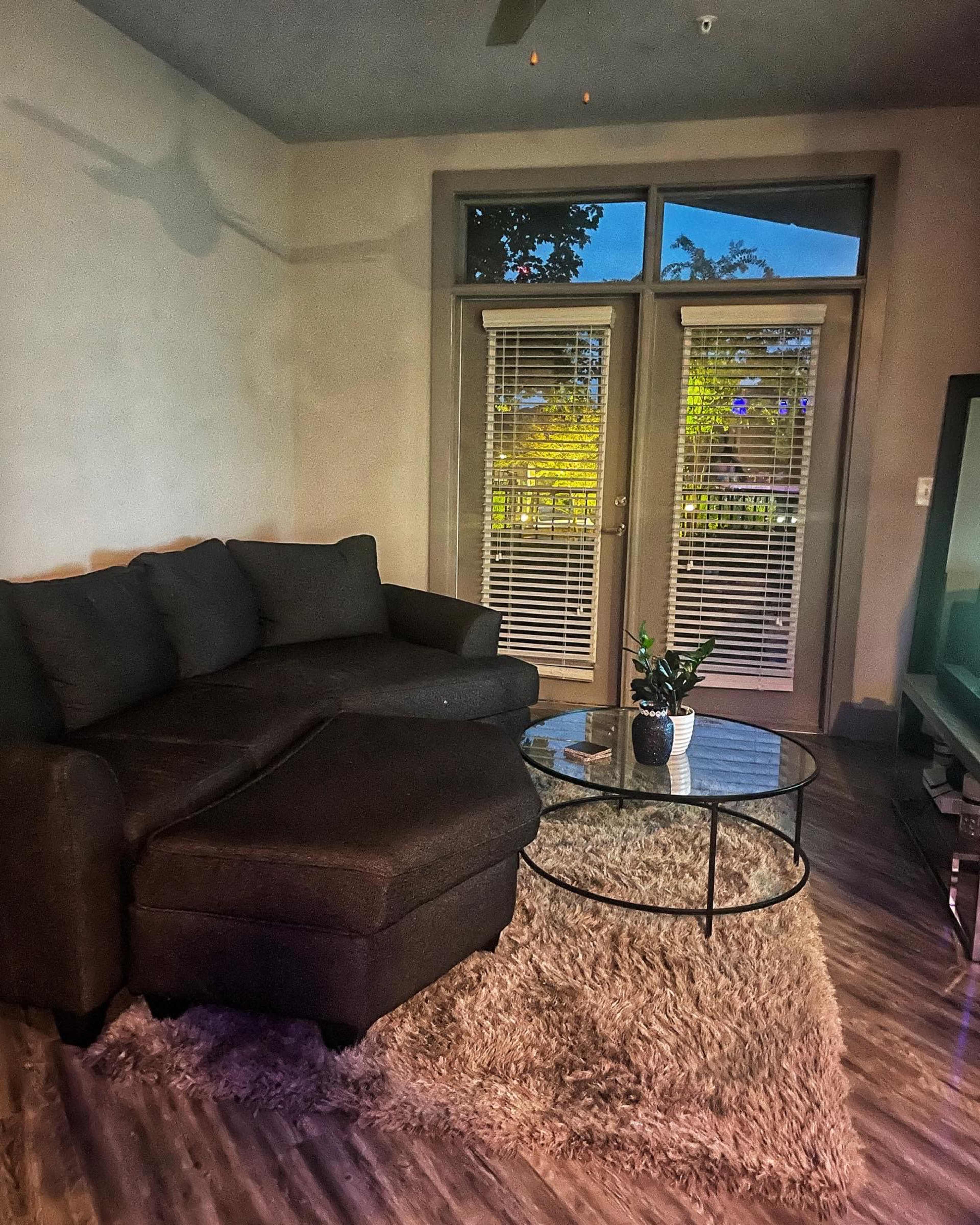 A dark sectional sofa and a glass coffee table sit on a rug in a well-lit living room with large sliding doors.