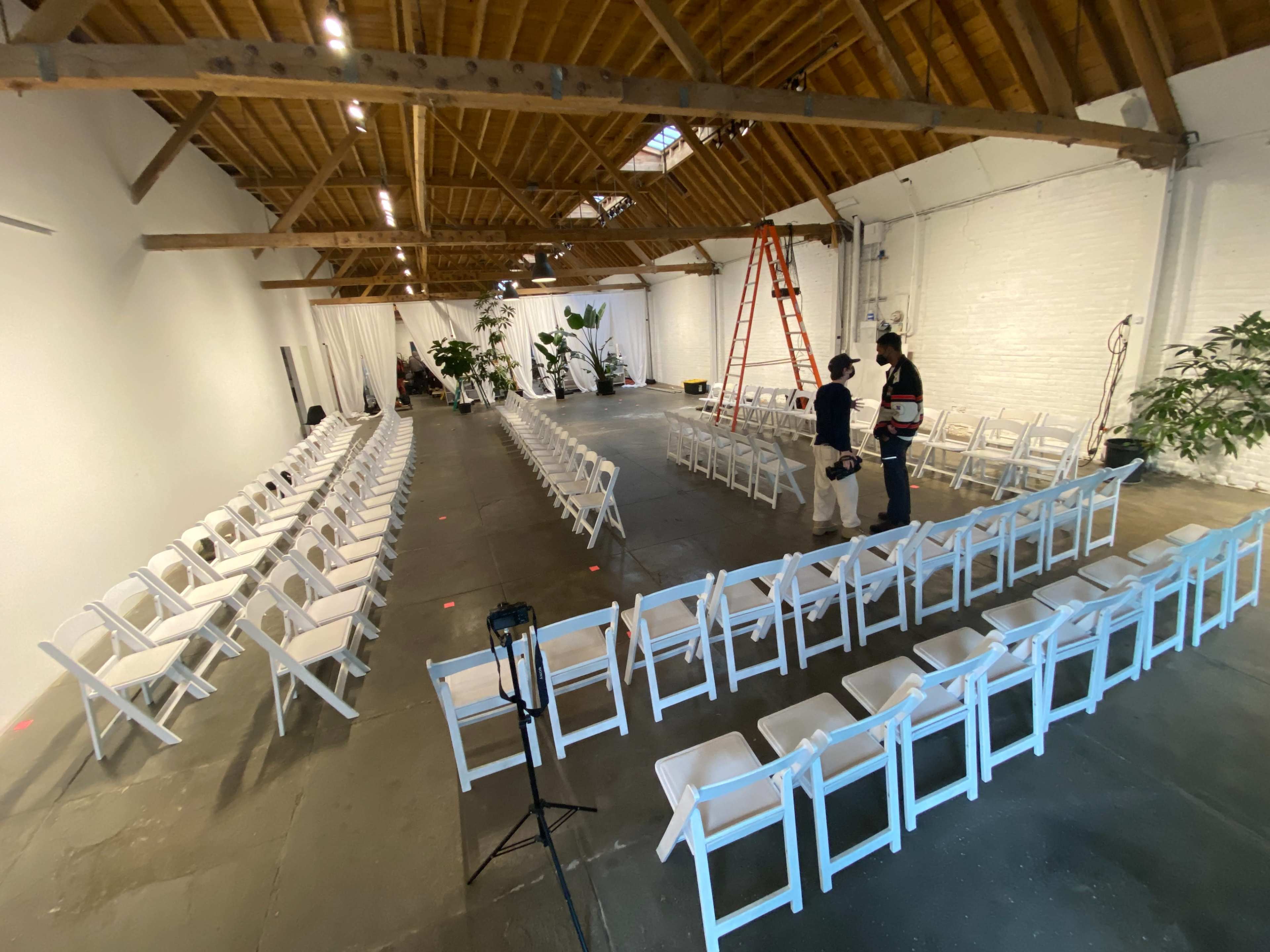 A spacious indoor venue features two rows of white chairs arranged in a semicircle, with a ladder and two individuals preparing the space.
