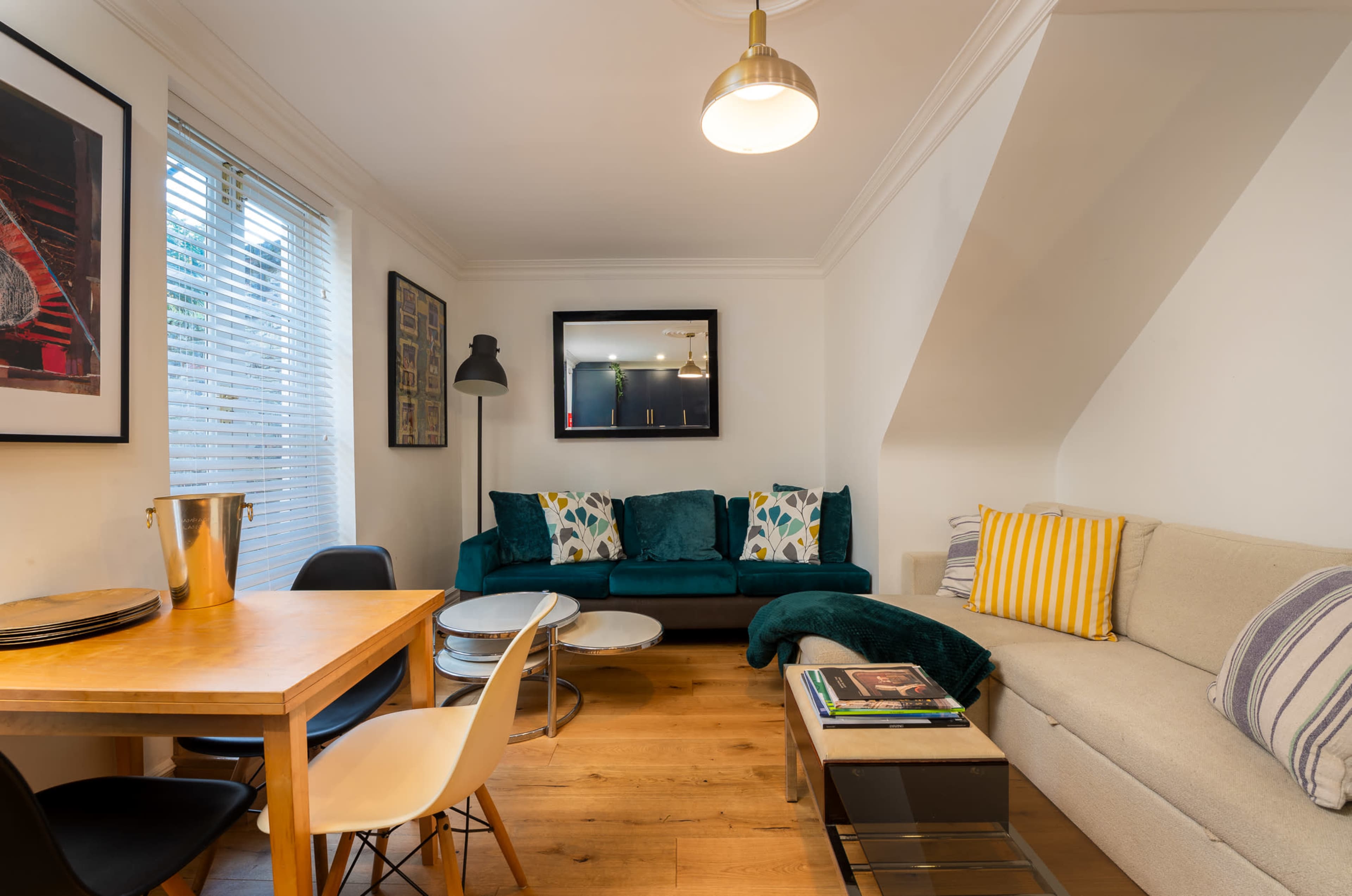 The image shows a cozy living space featuring a light wood dining table, a sofa with decorative cushions, and artwork on the walls.