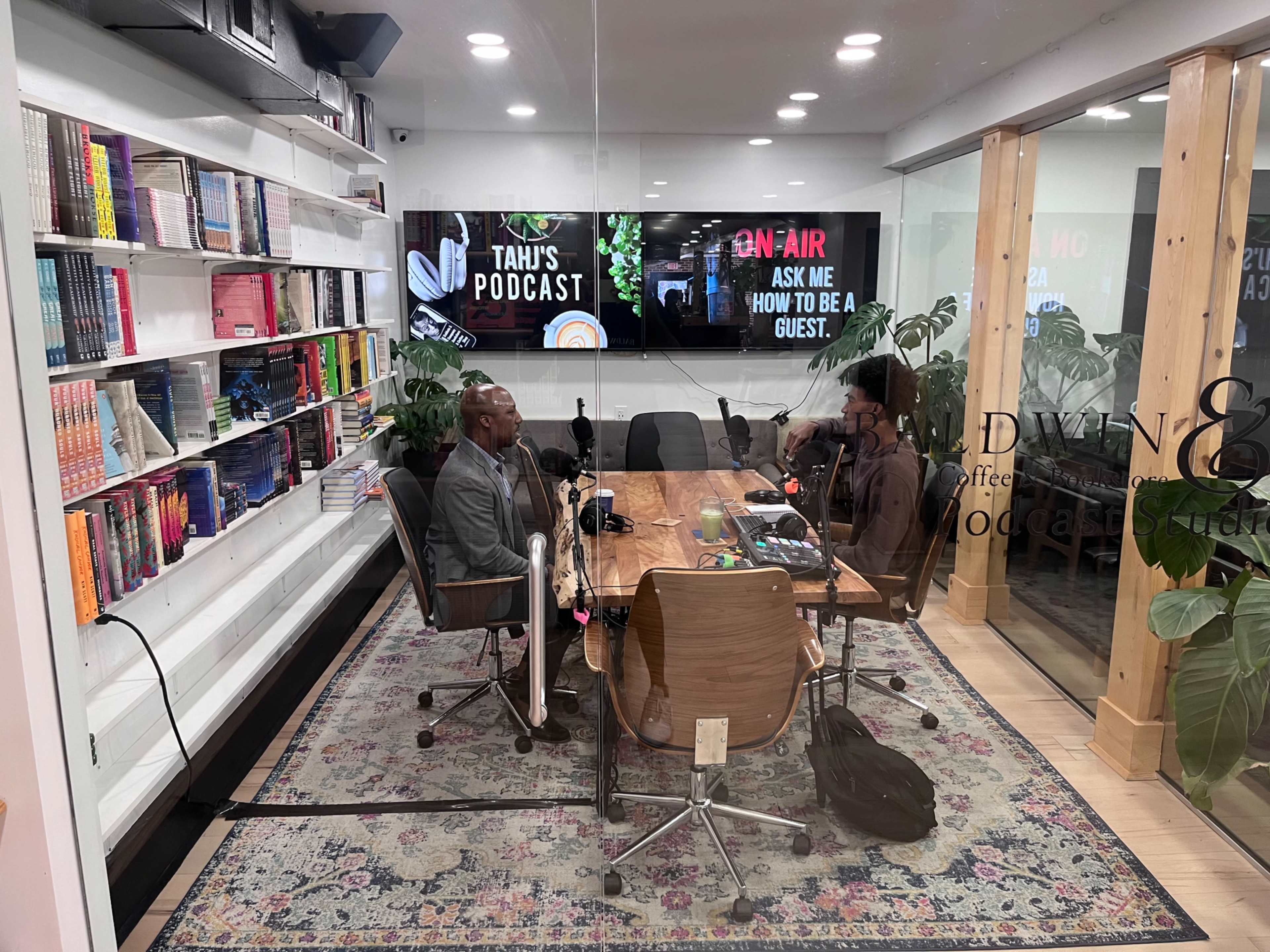 A podcast recording studio features a wooden table with microphones and two seated guests, surrounded by bookshelves and a large screen displaying "Tahu's Podcast."