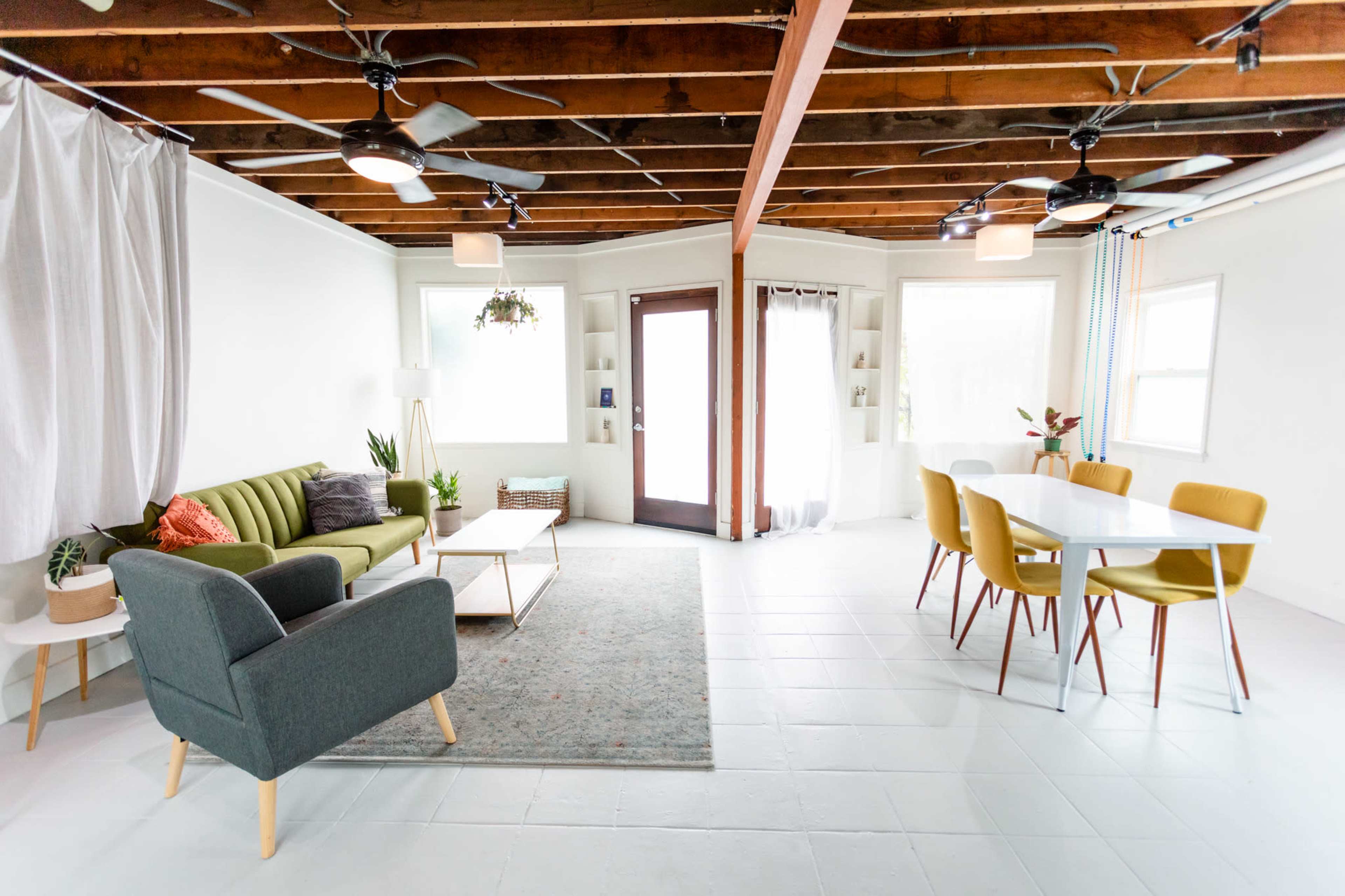 The image shows a bright and airy living room with a green sofa, a gray armchair, a coffee table, and a dining area featuring a white table and yellow chairs under wooden beams.