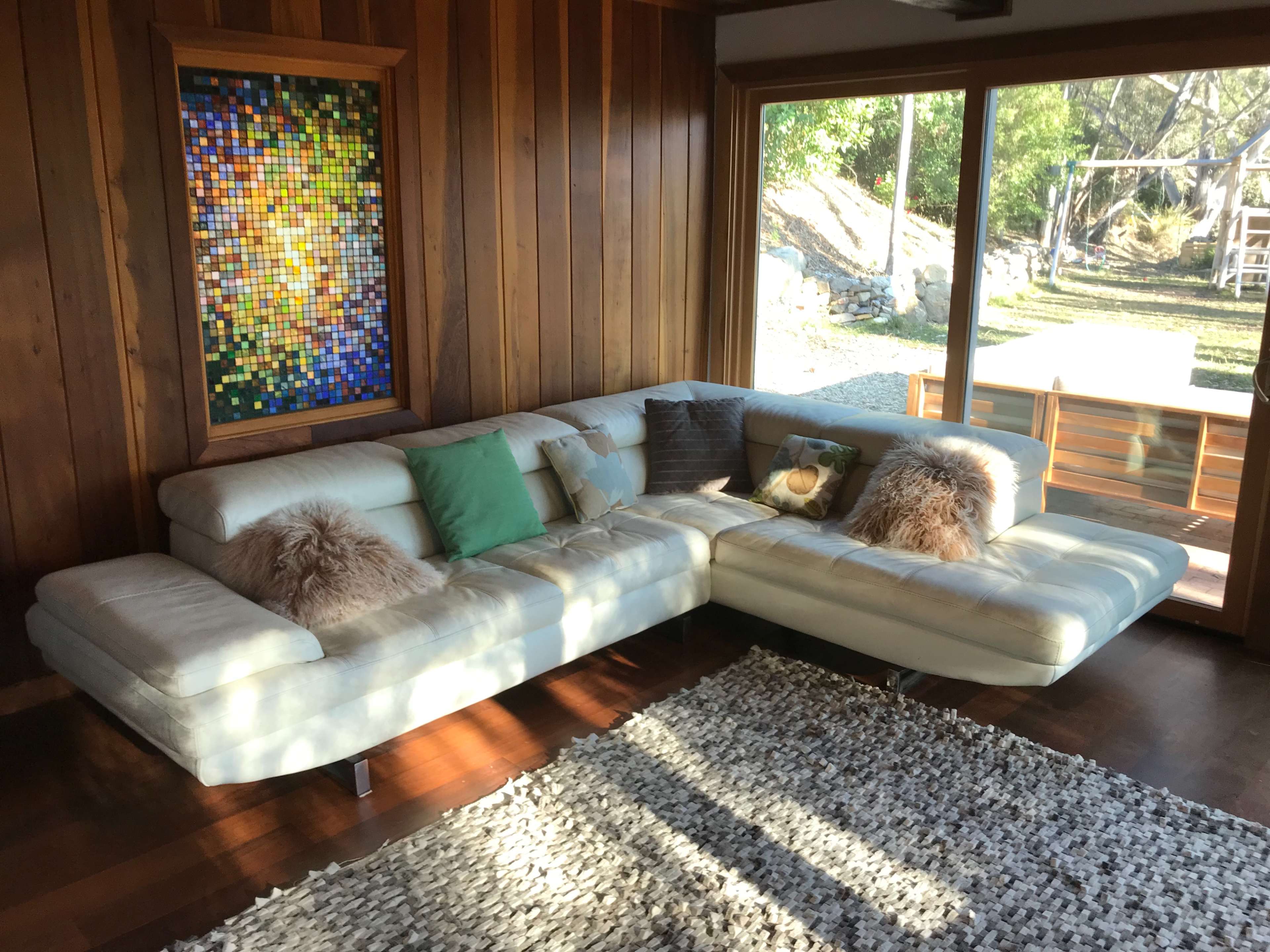 A light-colored sectional sofa with decorative pillows is positioned in a room featuring wooden walls and a large window overlooking a grassy area.