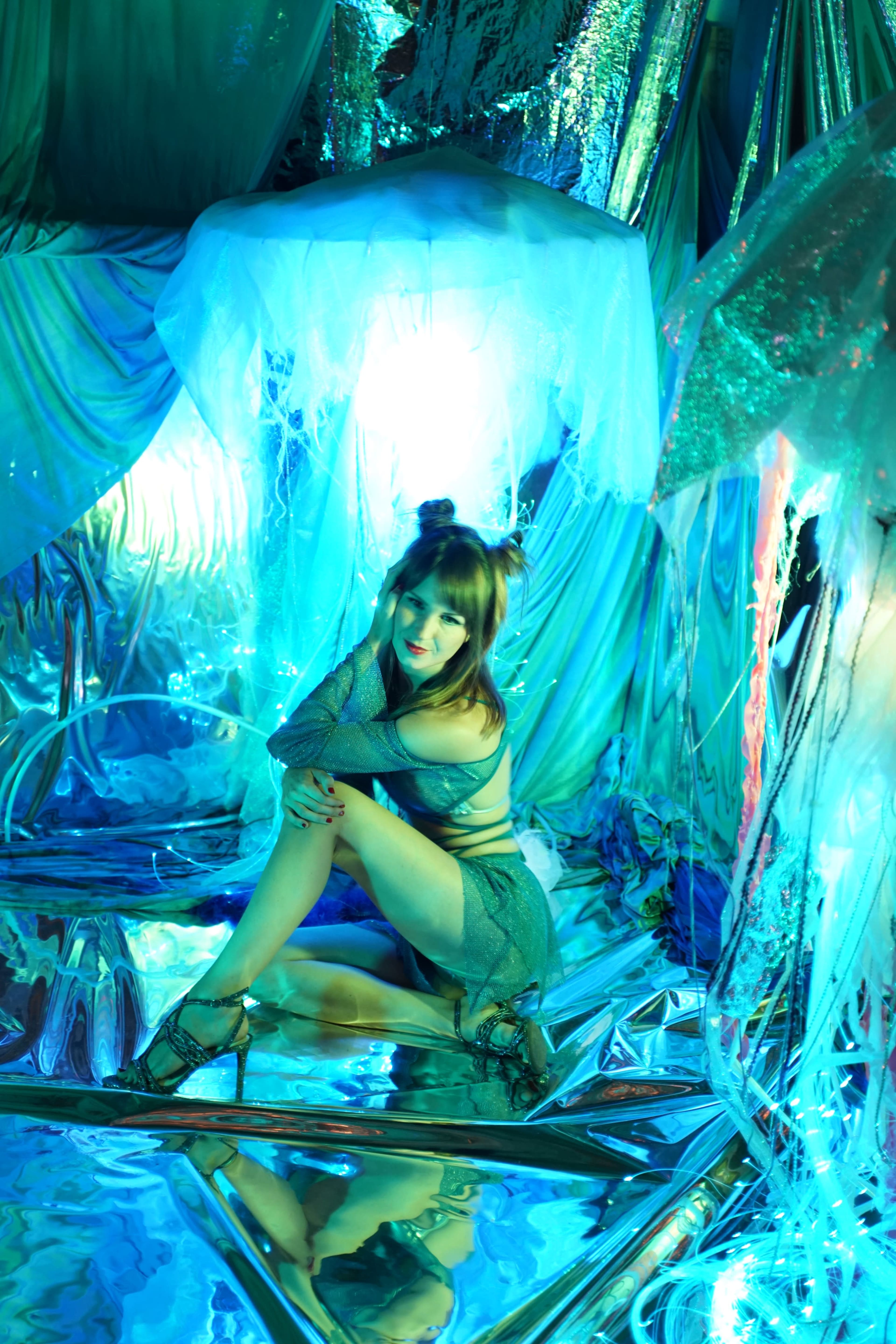 A person sits on a reflective surface surrounded by shimmering blue and silver fabric in a creatively designed space.