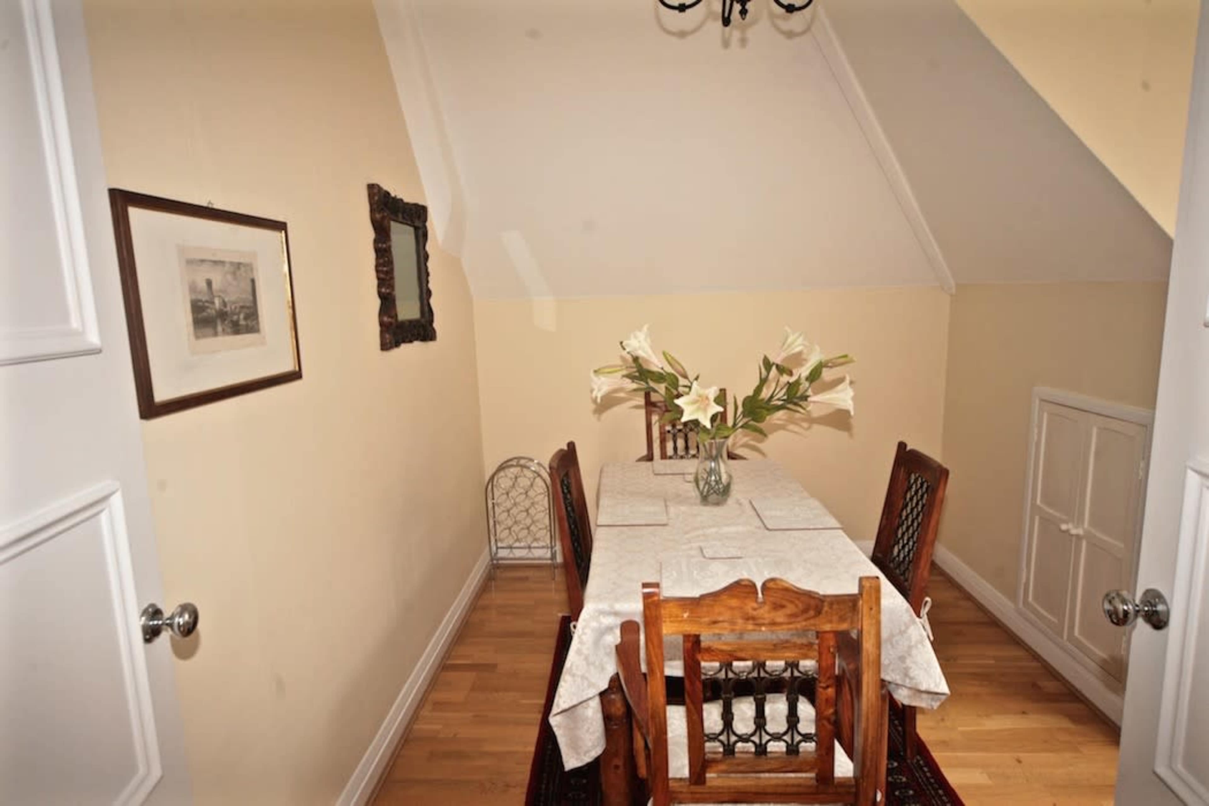 A small dining area features a wooden table surrounded by four chairs, with a vase of lilies on the table, and walls adorned with framed pictures.