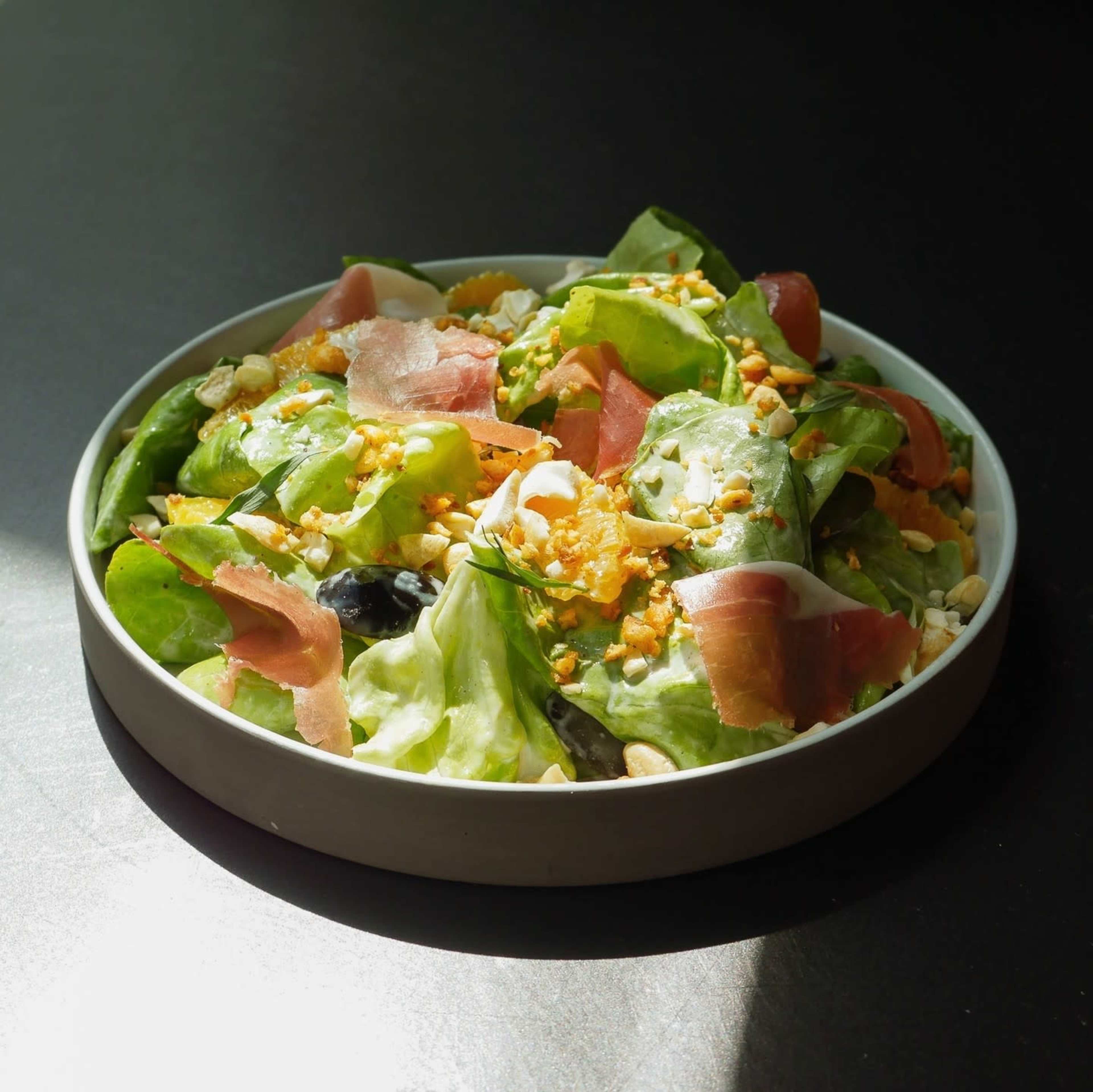 A serving of salad with greens, ham, nuts, and a creamy dressing is displayed in a shallow bowl on a dark surface.