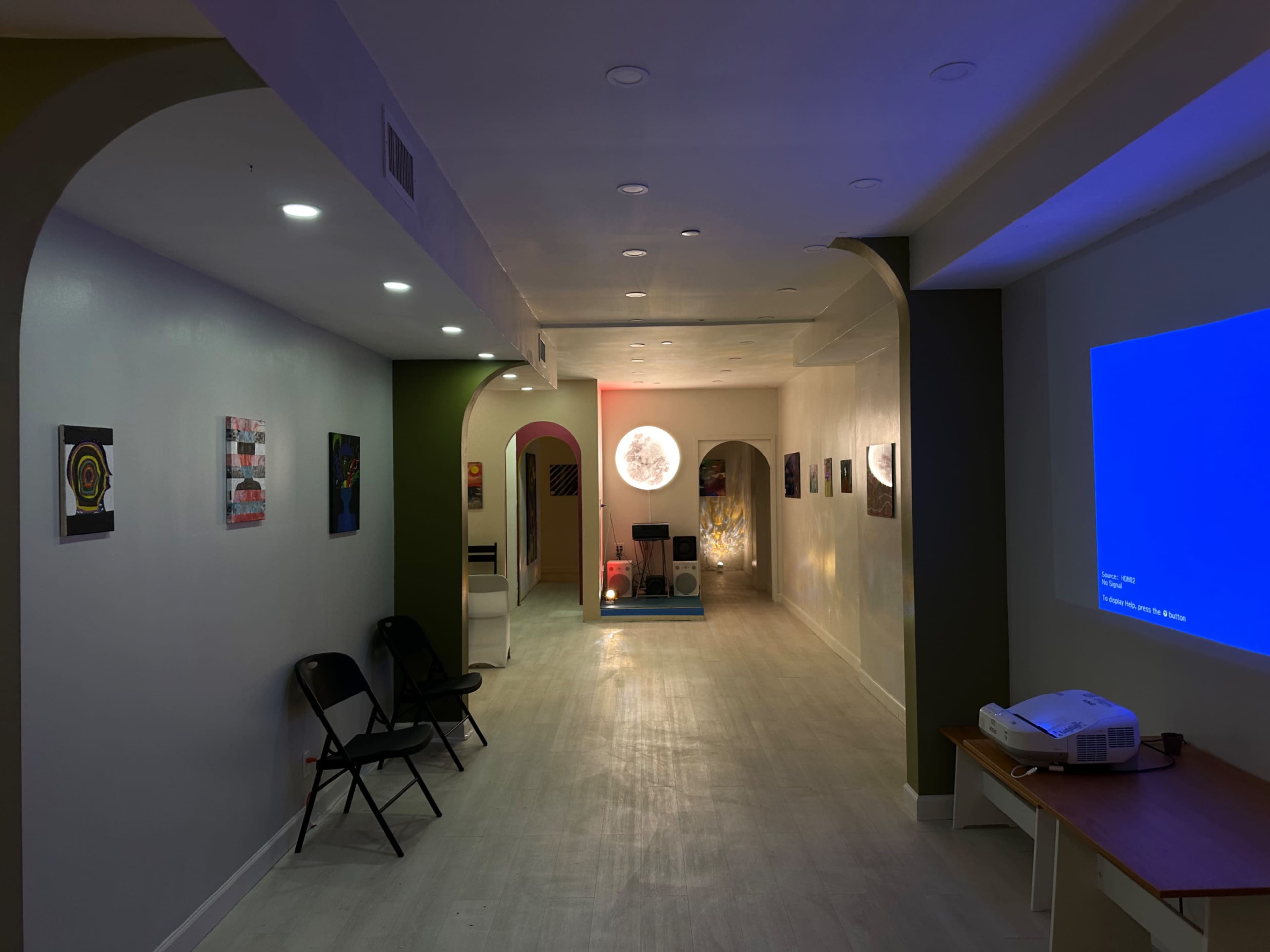 The image shows a long, narrow corridor with white walls, archways, colorful artwork displayed, and a projector casting a blue light on one wall.