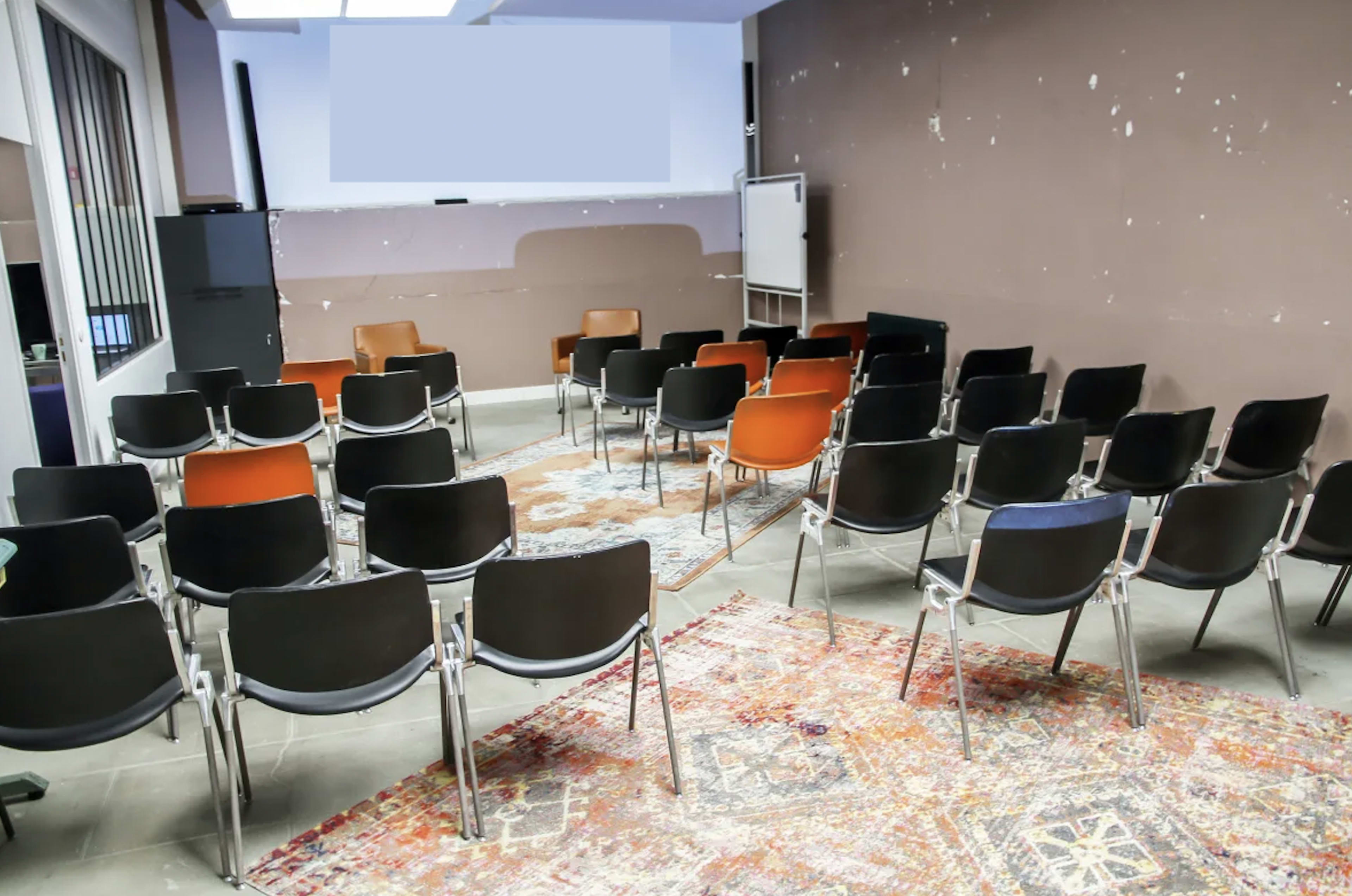 A room with a mix of black and orange chairs arranged in a semi-circle around a central space, with a patterned rug on the floor and a wall displaying some wear.