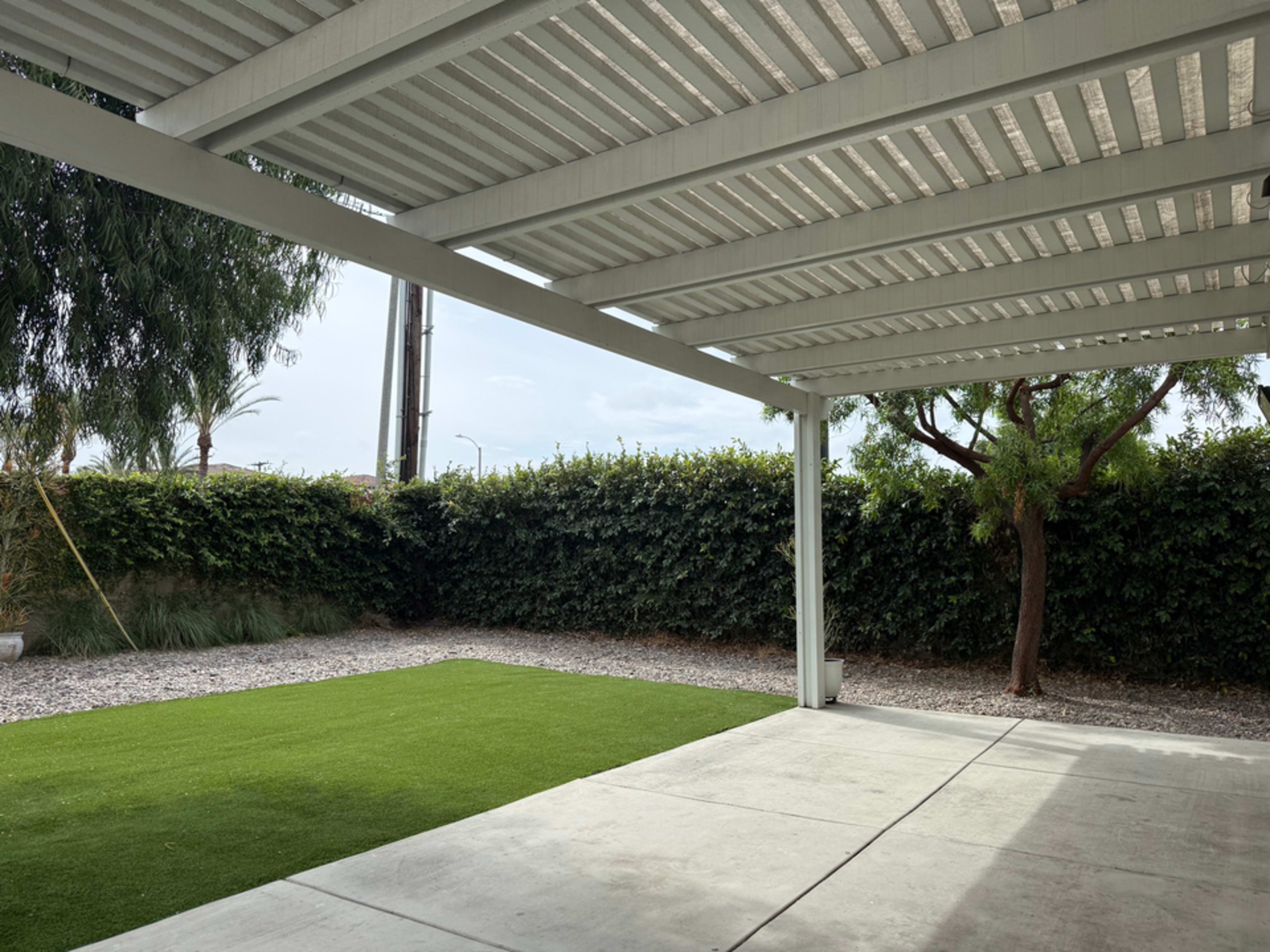 The image shows a shaded patio area with a concrete floor and artificial grass, bordered by a low hedge and trees.