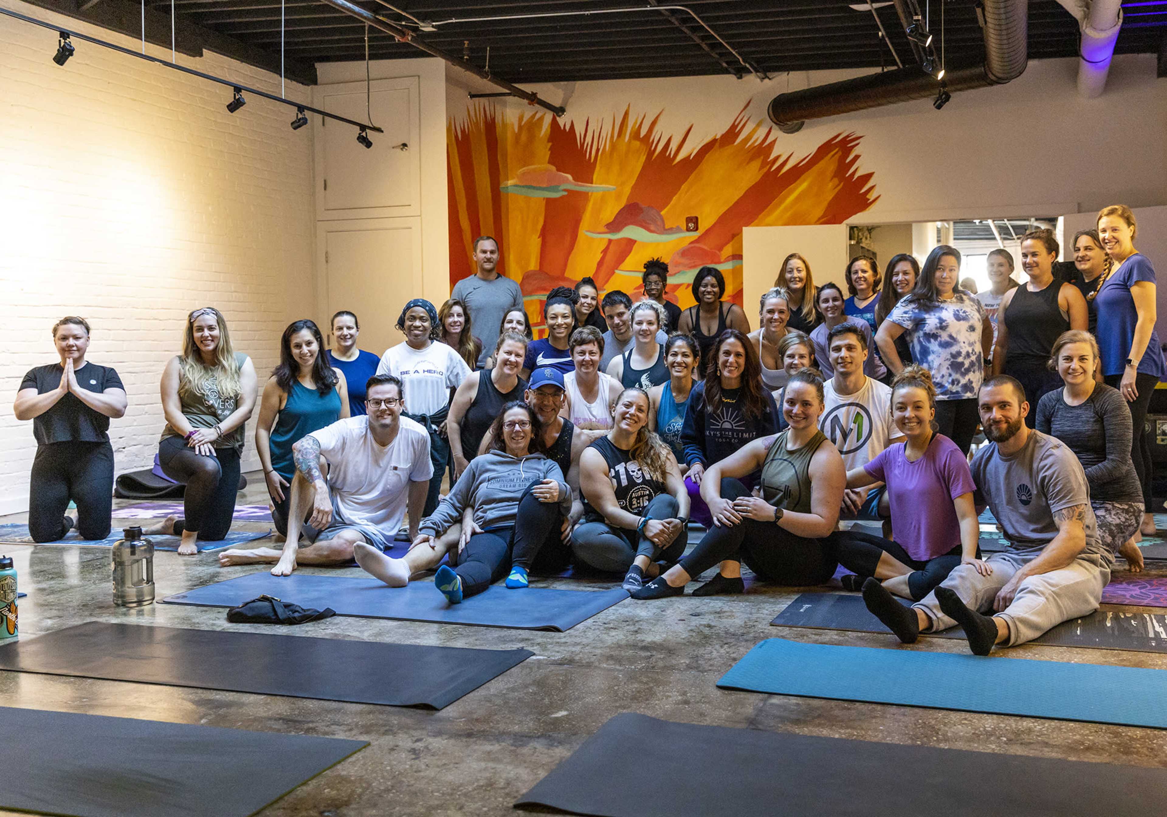 A large group of people is gathered on yoga mats in a studio with a colorful mural in the background.