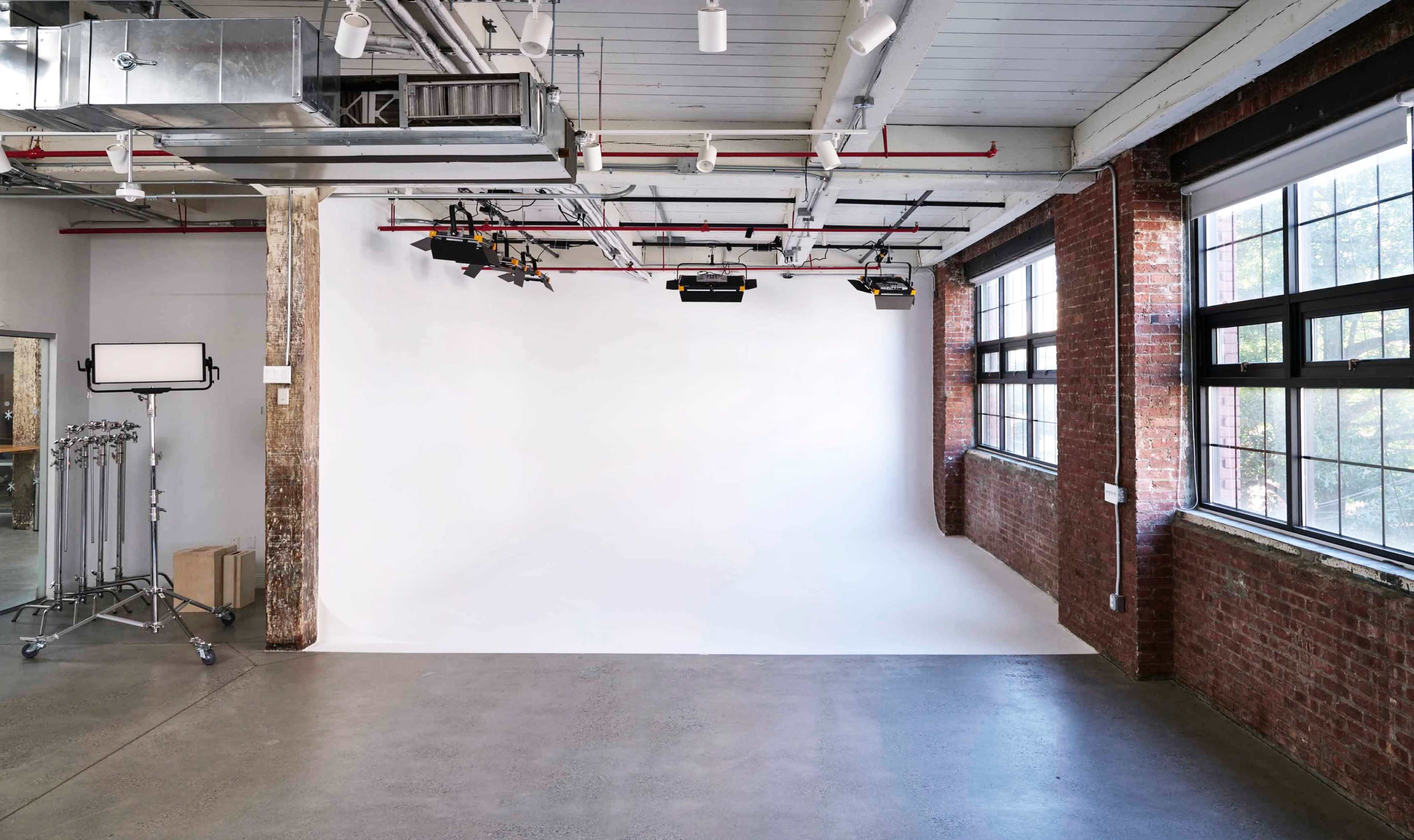 A spacious studio with a white backdrop and large windows, featuring exposed brick walls and industrial lighting fixtures.