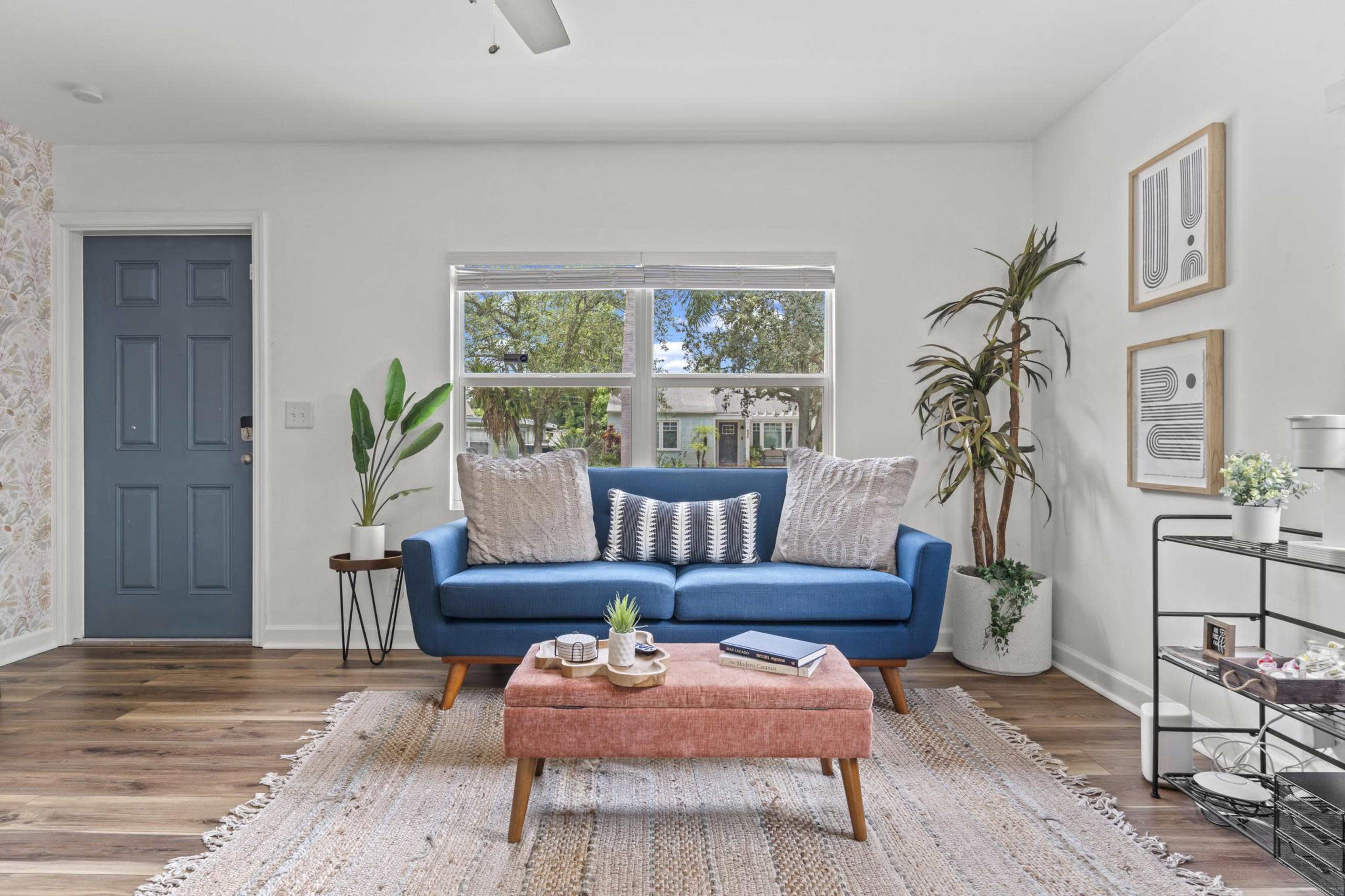 A bright living room features a blue couch with decorative pillows, a wooden coffee table, and large windows overlooking greenery.
