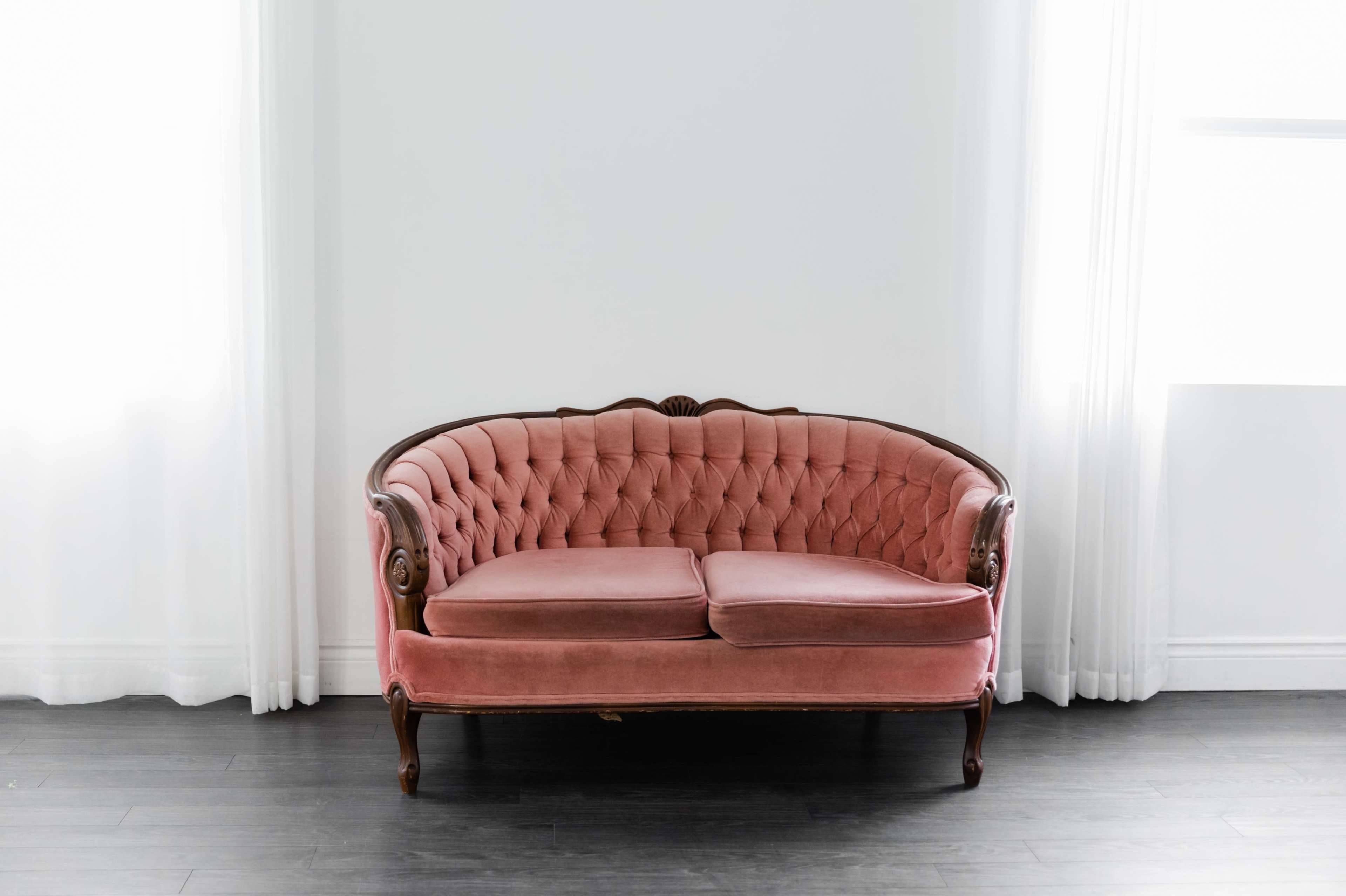 A pink tufted sofa sits in a brightly lit room with white curtains and dark wood flooring.