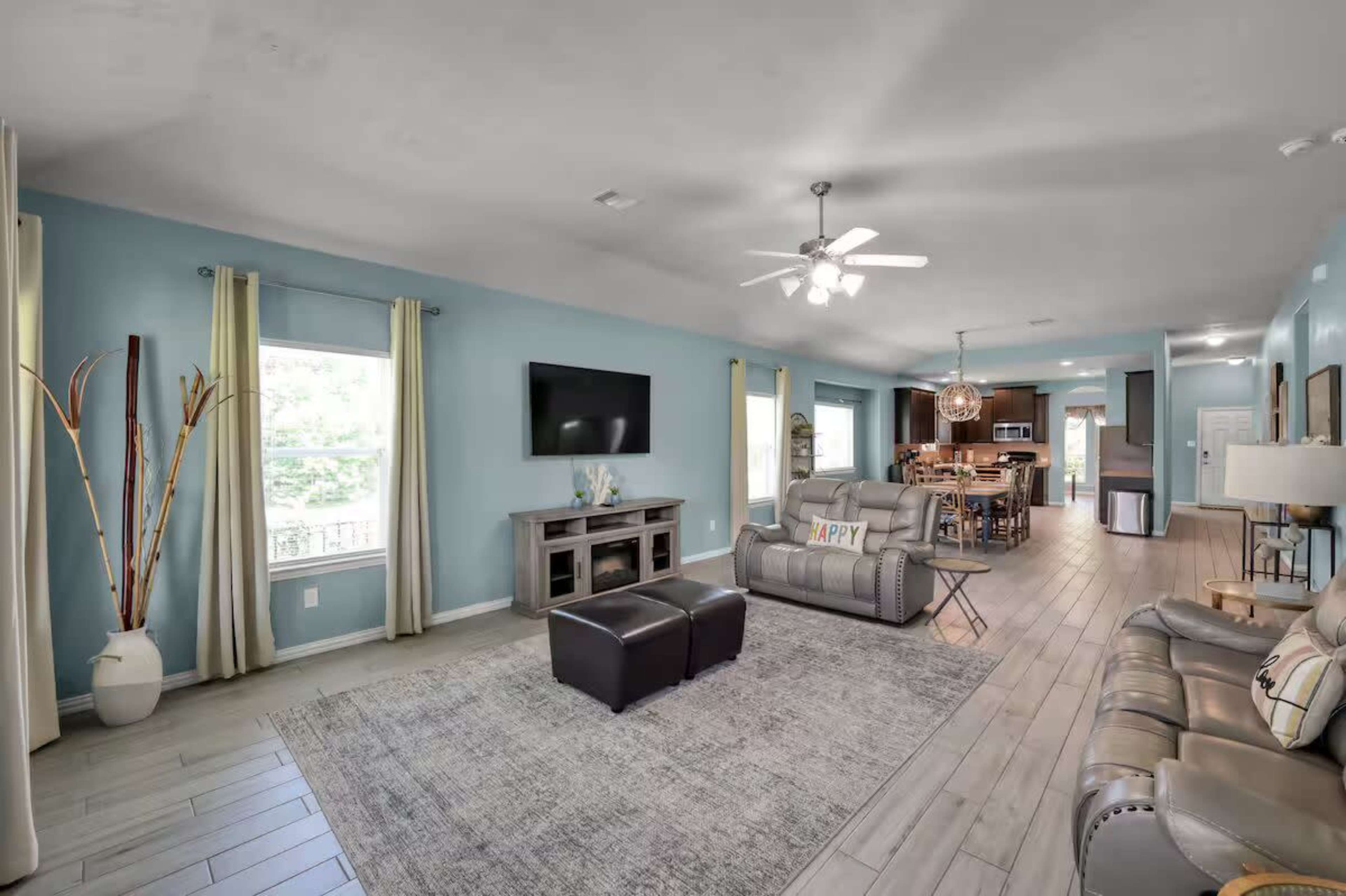 The image depicts a spacious living room with light blue walls, a television mounted on the wall, a light fixture above, and a view into a kitchen area in the background.