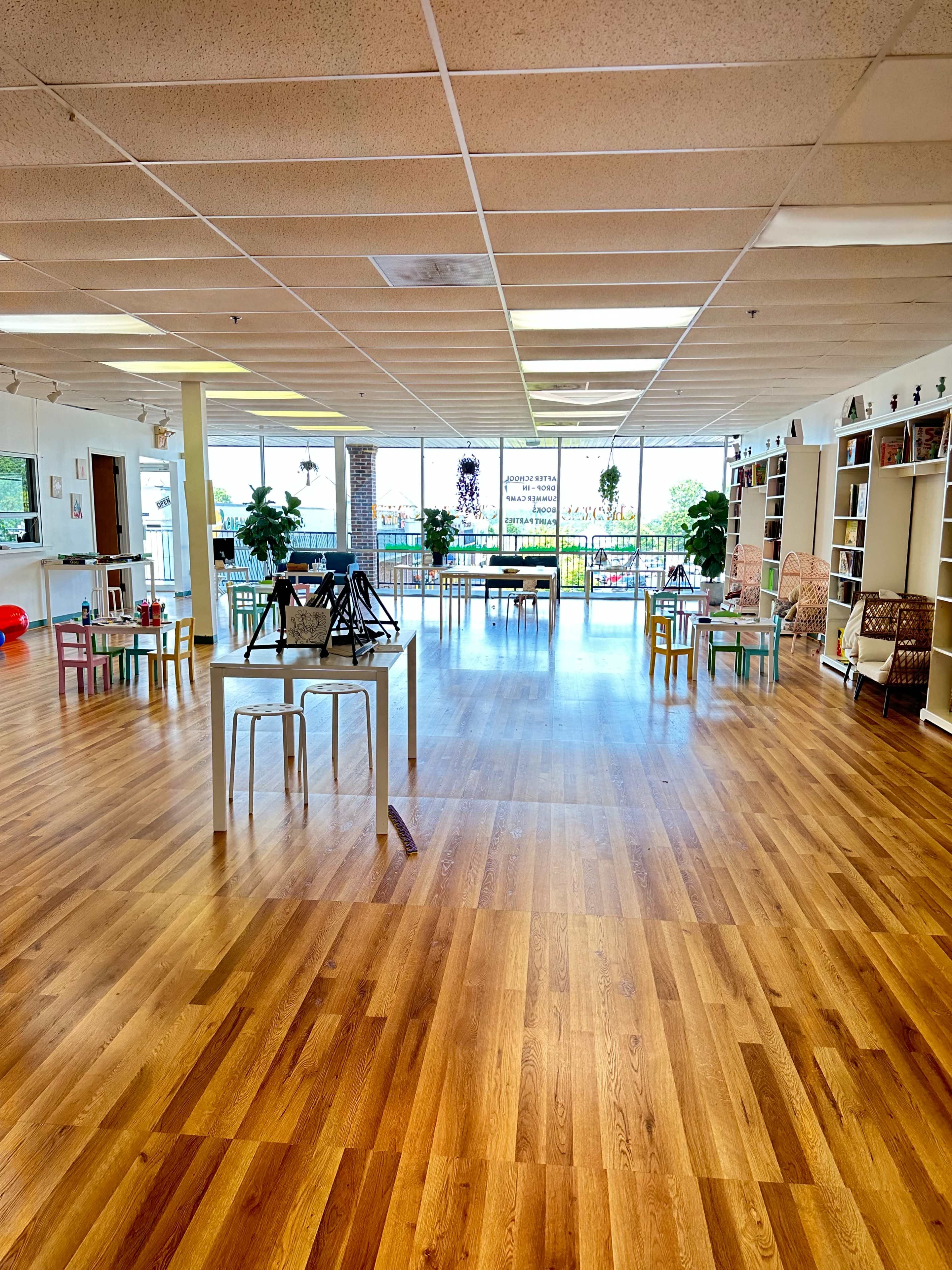 The image shows a spacious room with wooden floors, several tables surrounded by chairs, and large windows letting in natural light.