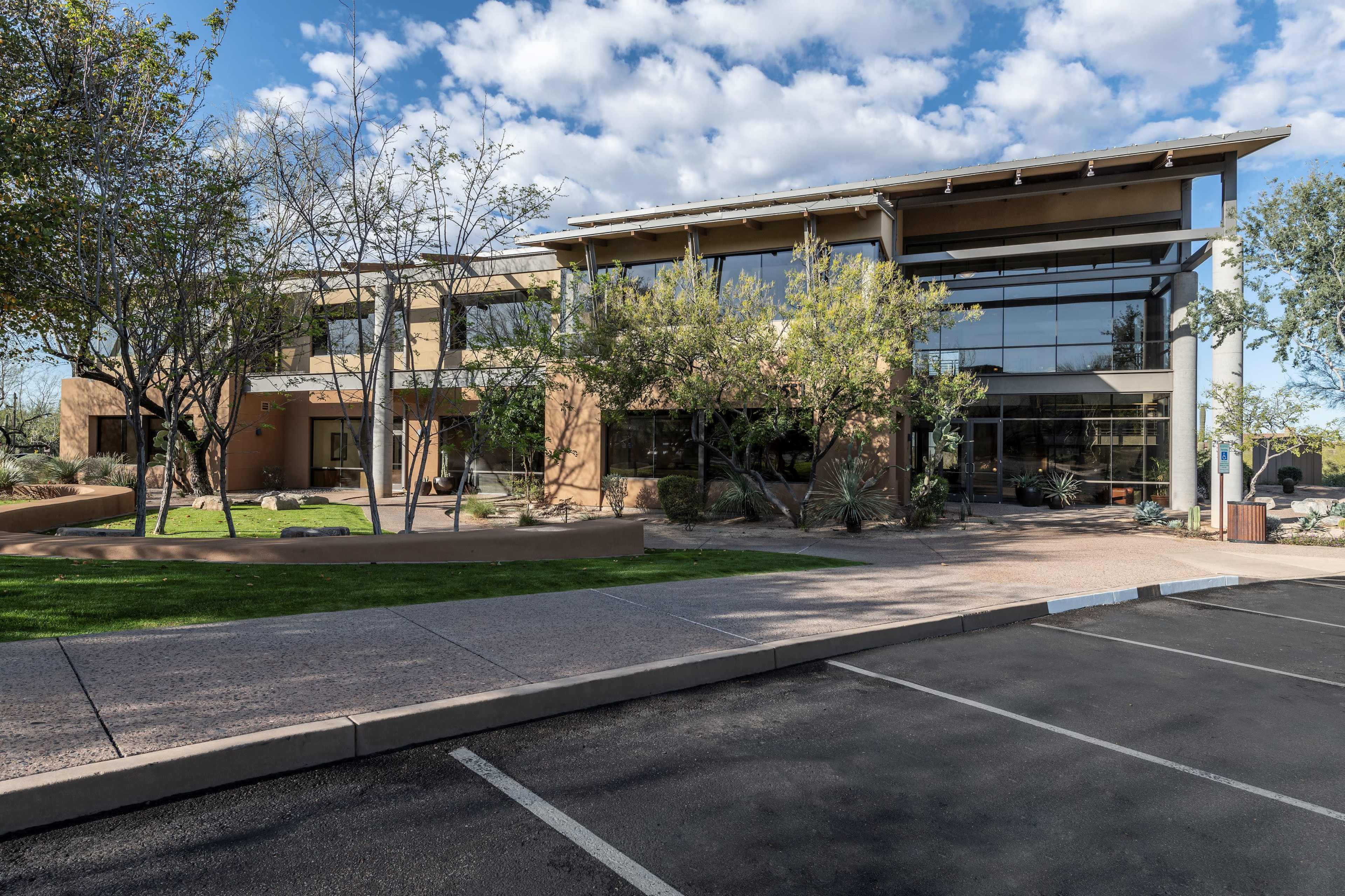 The image features a modern two-story office building surrounded by trees and shrubs, with a parking lot in the foreground.