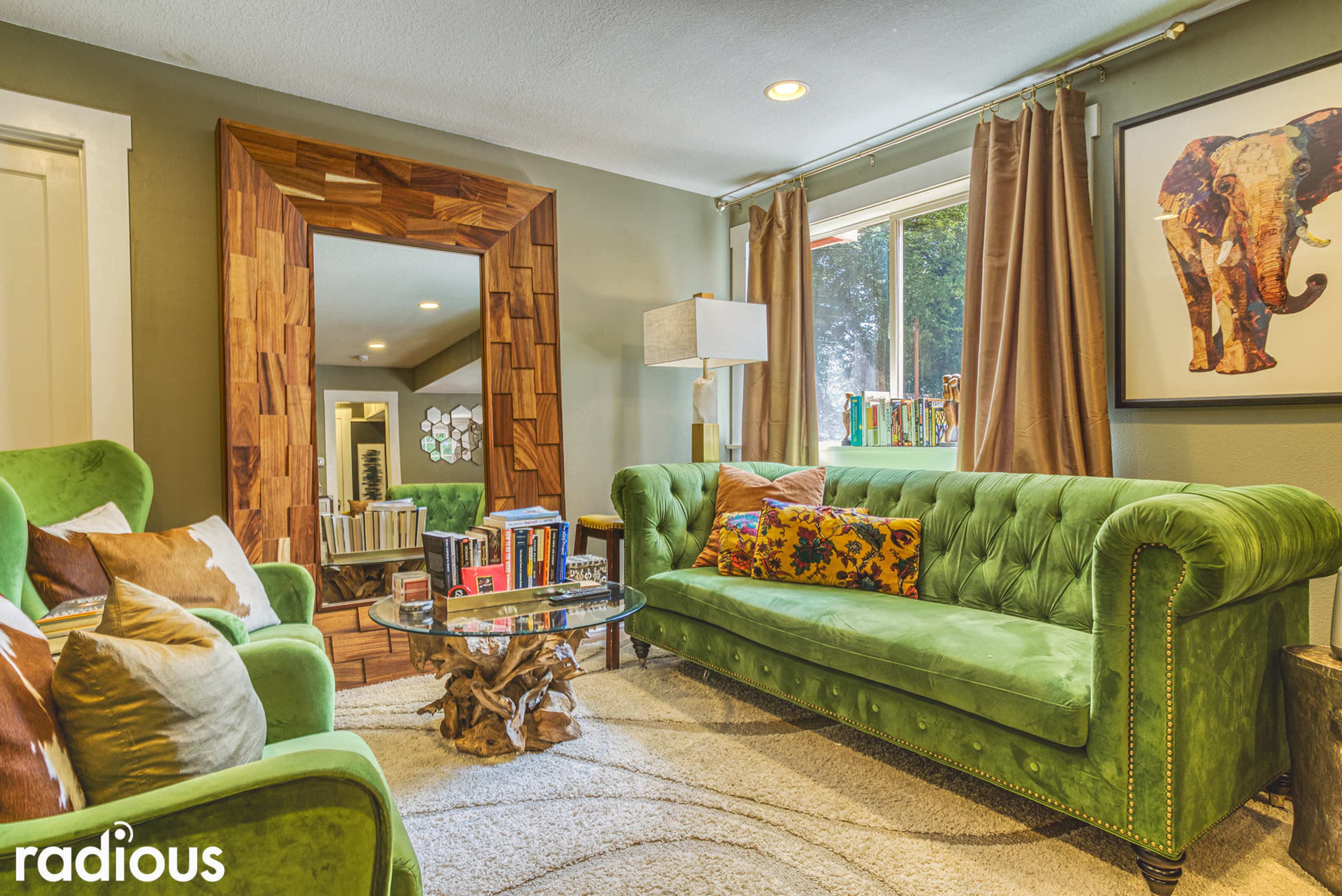 A cozy living room features a green velvet sofa, a round glass coffee table, and a large rectangular mirror framed with wood.