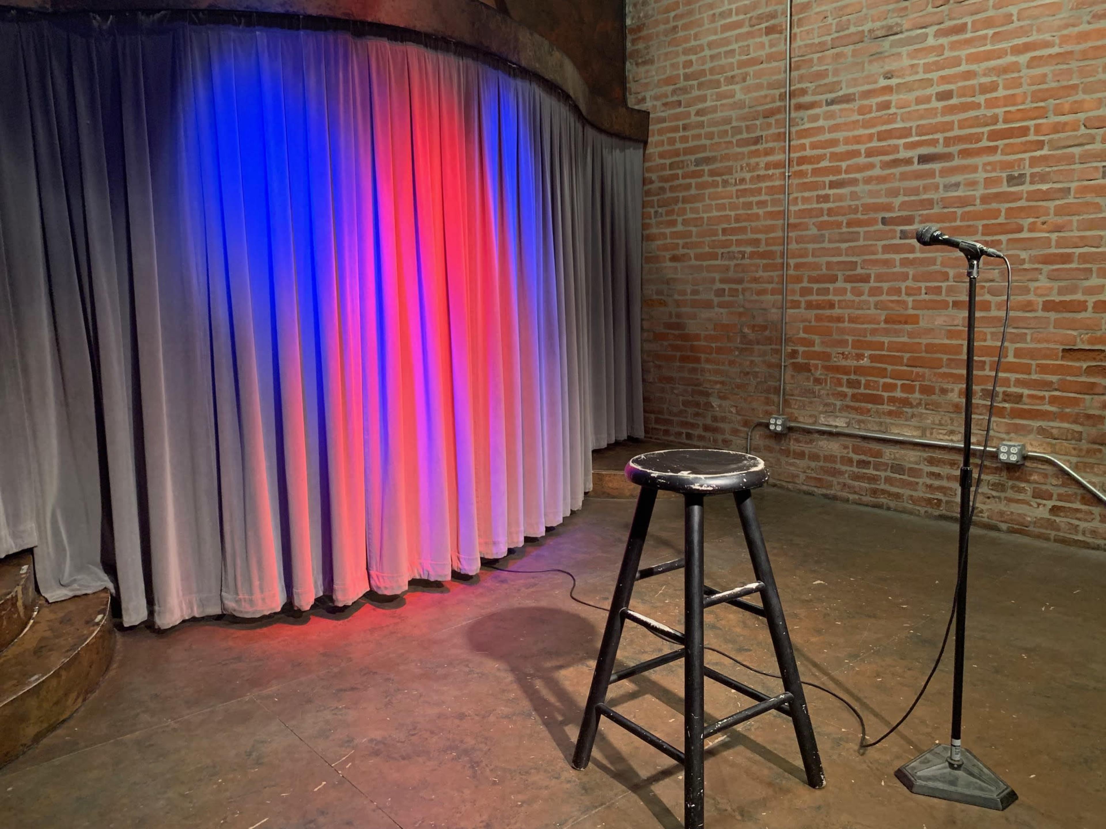 A black stool and microphone stand are positioned in front of a backdrop of draped gray curtains illuminated with red and blue lights in a brick-walled room.