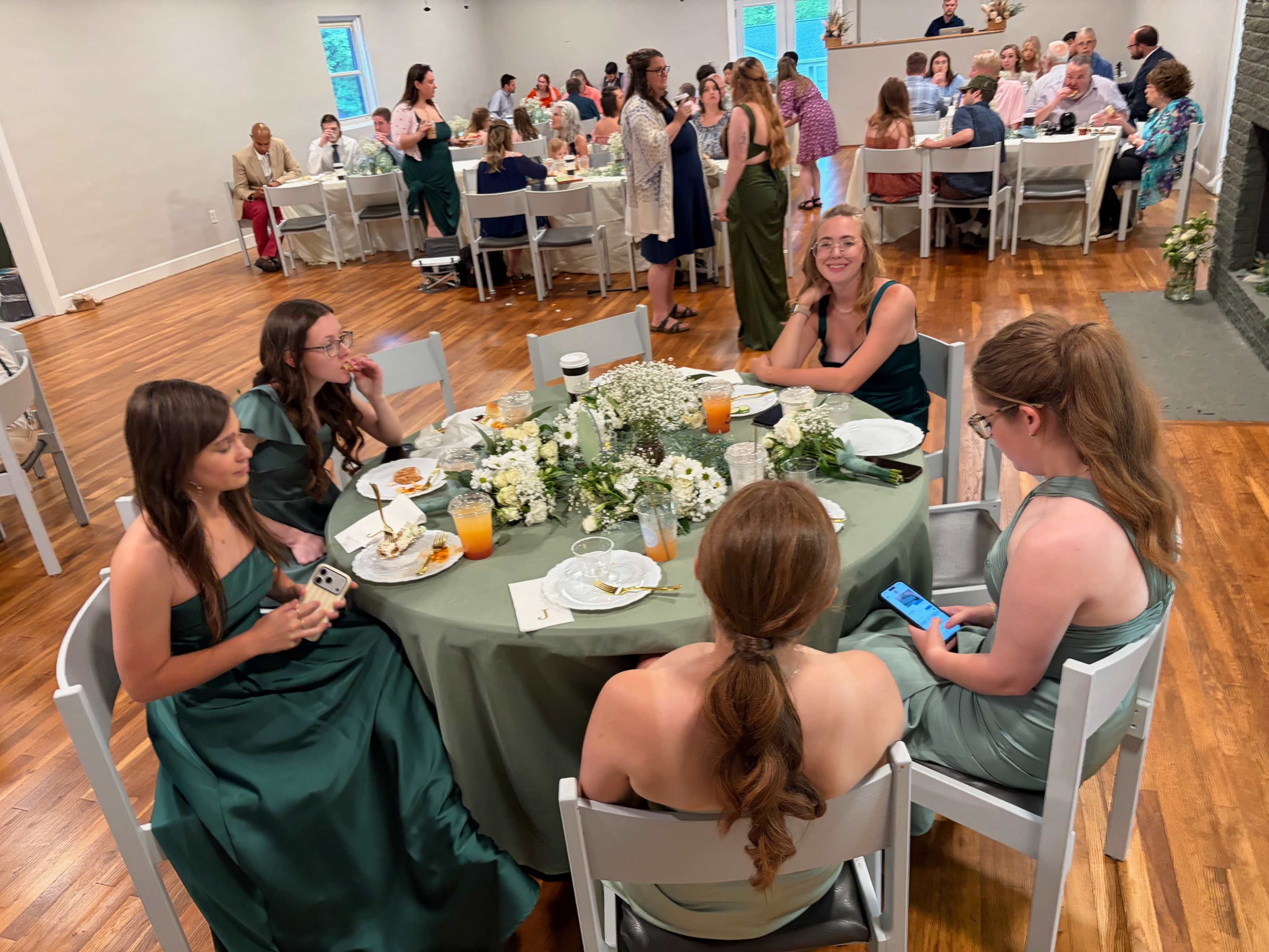 A group of women in green dresses sit around a table adorned with floral centerpieces at a lively event with many guests in the background.