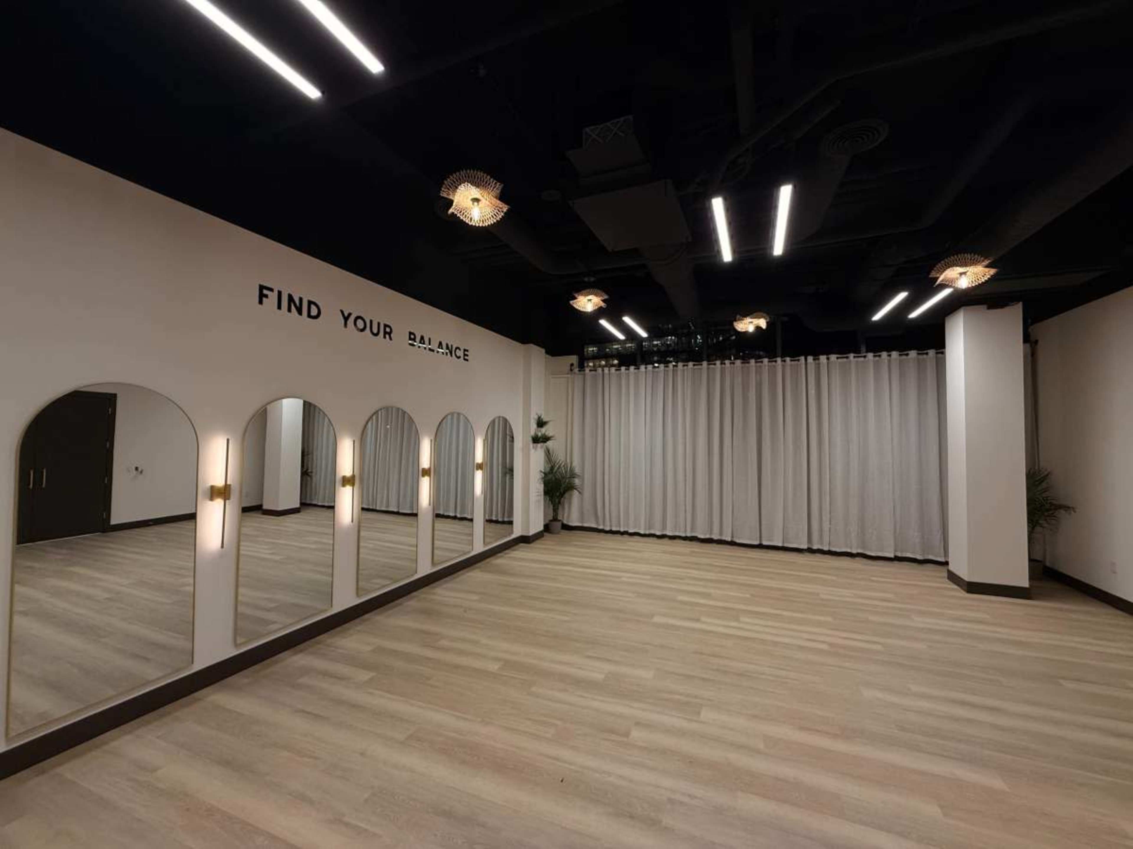 The image shows a spacious studio with a light wooden floor, large mirror arches on one wall, and soft lighting fixtures hanging from the ceiling.